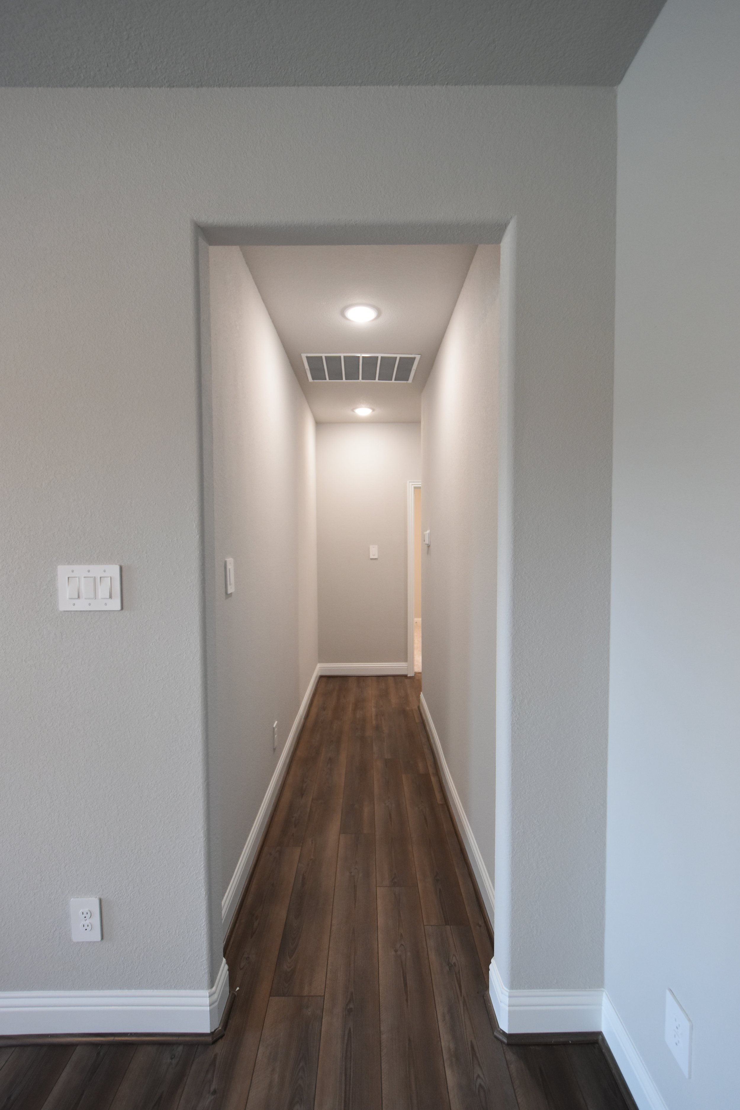 Empty hallway with white walls, dark wood flooring, ceiling lights, air vent, and several electrical outlets and switches.