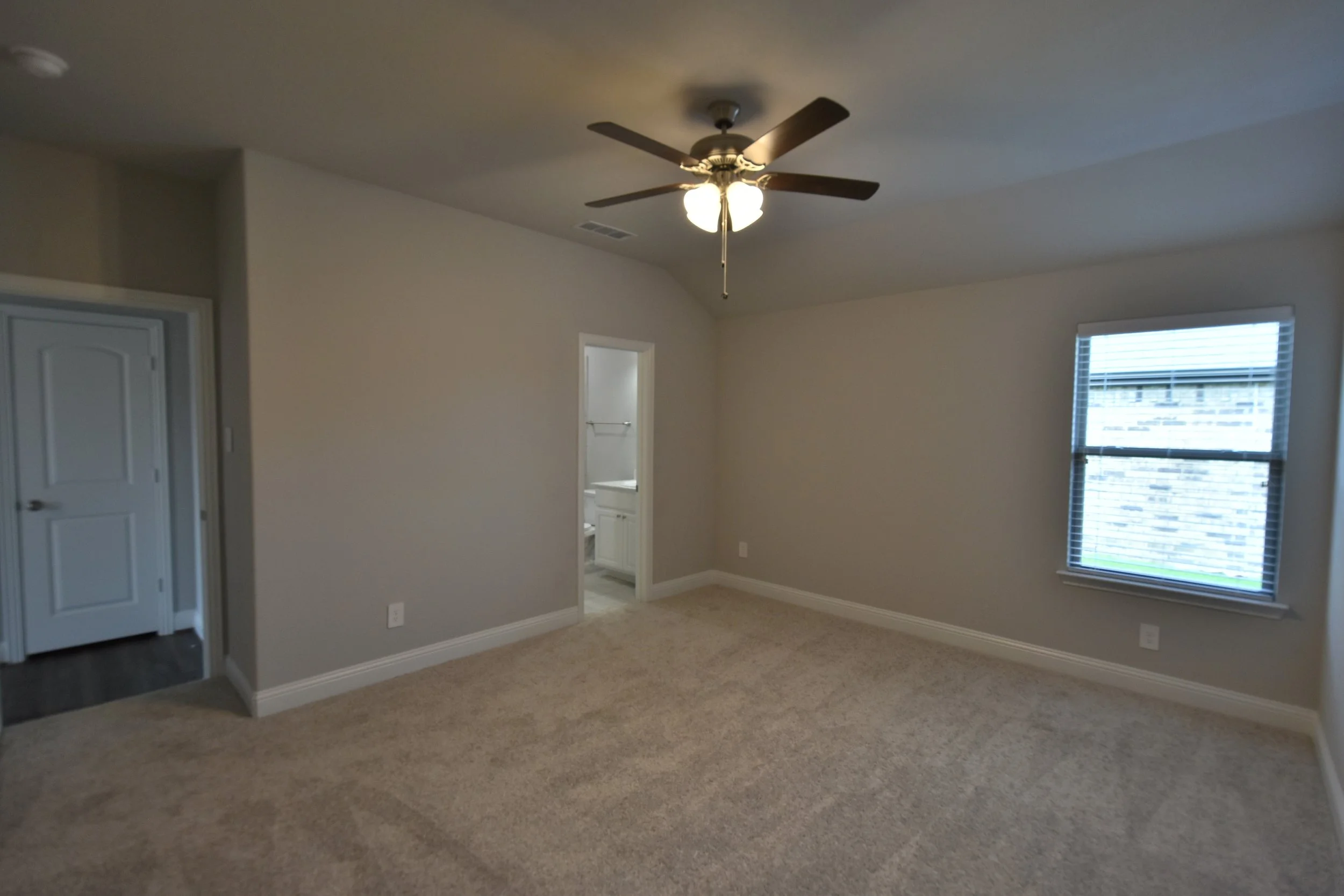 Empty room with beige carpet, white walls, ceiling fan with lights, two windows with blinds, and a doorway leading to a bathroom.