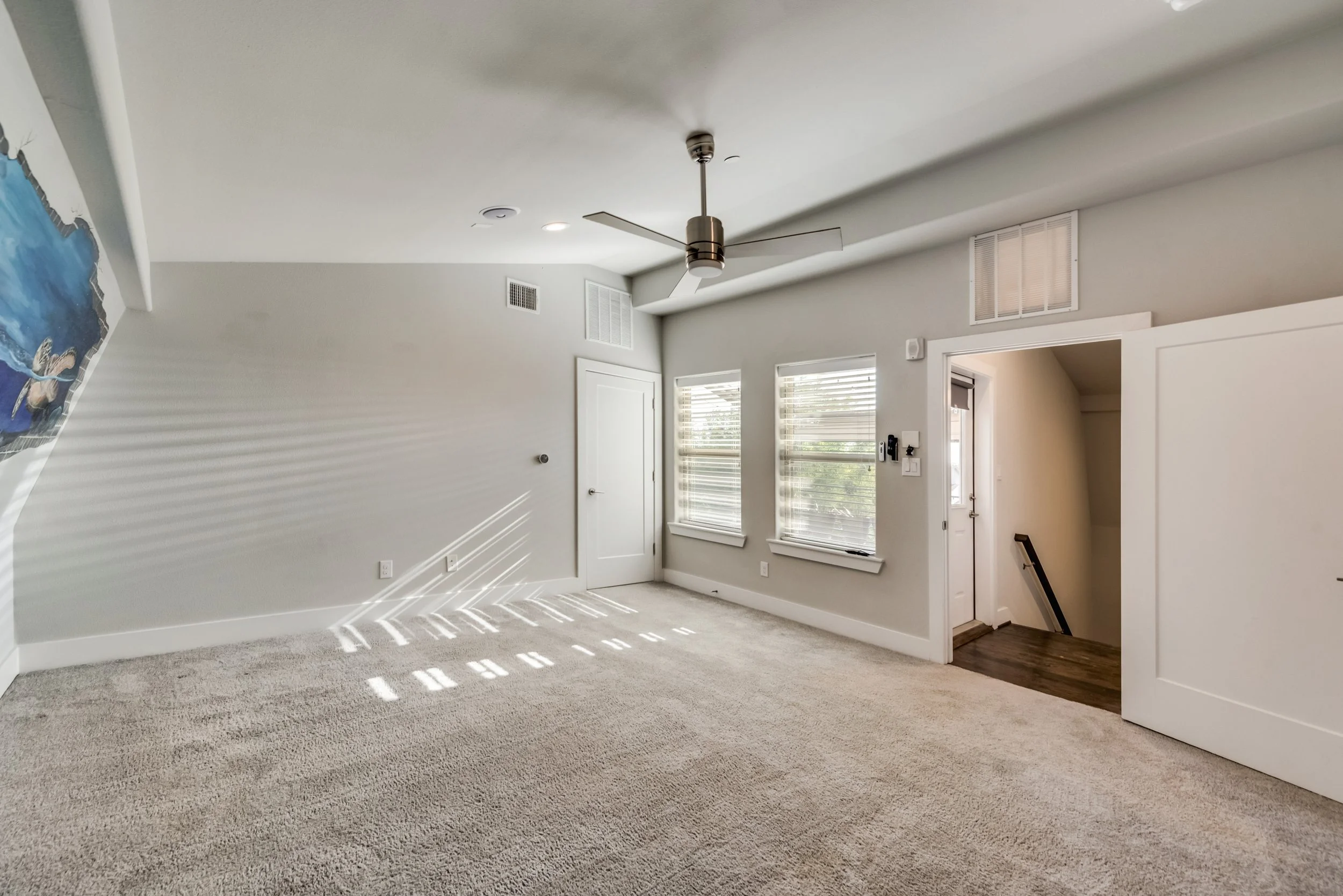 Empty living room with beige carpet, white walls, windows with blinds, a ceiling fan, and an open doorway leading to a staircase.