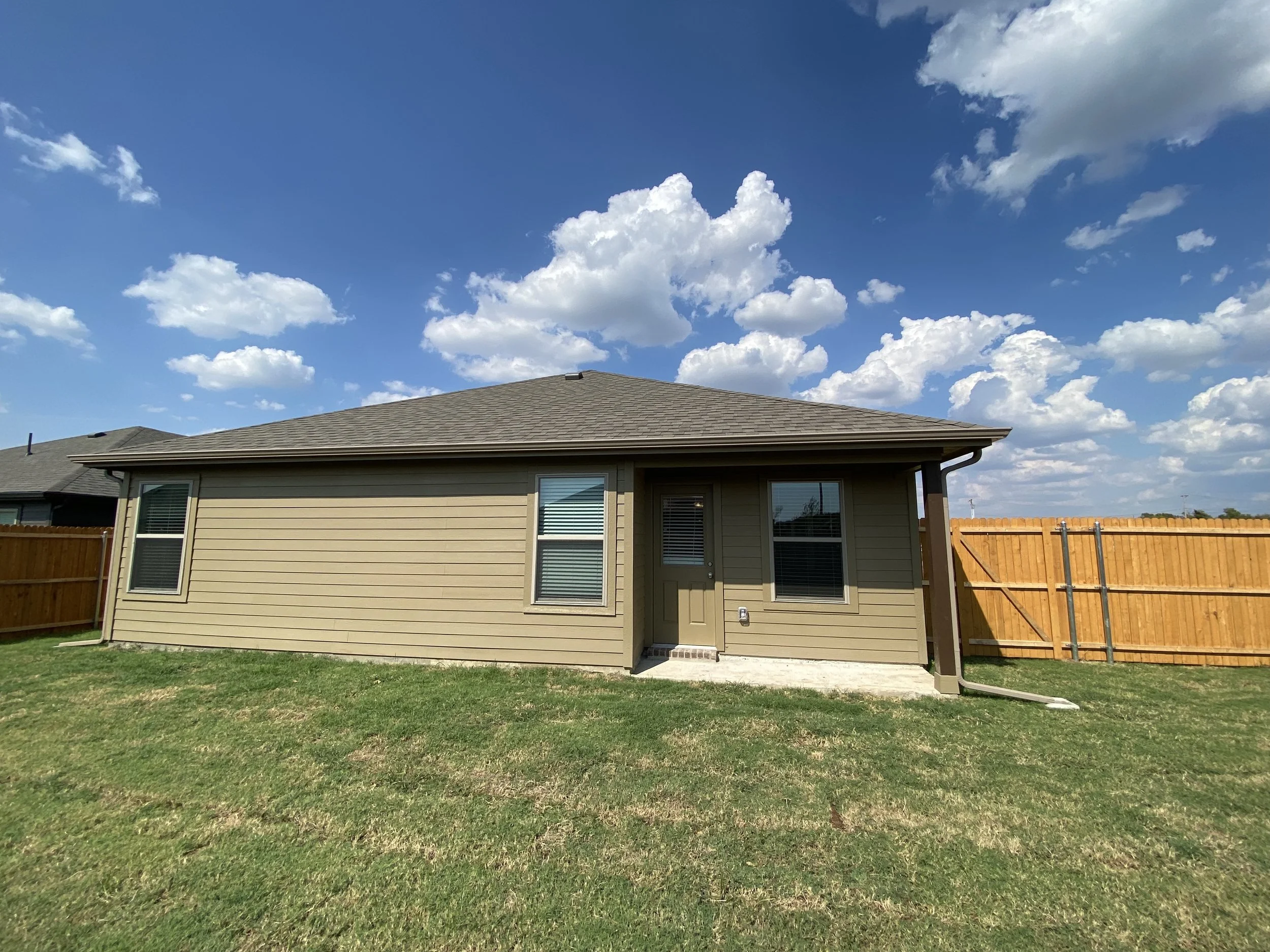 The image shows the back of a beige house with white-framed windows and a door, a green lawn, and a wooden fence, under a mostly clear blue sky with some fluffy white clouds.