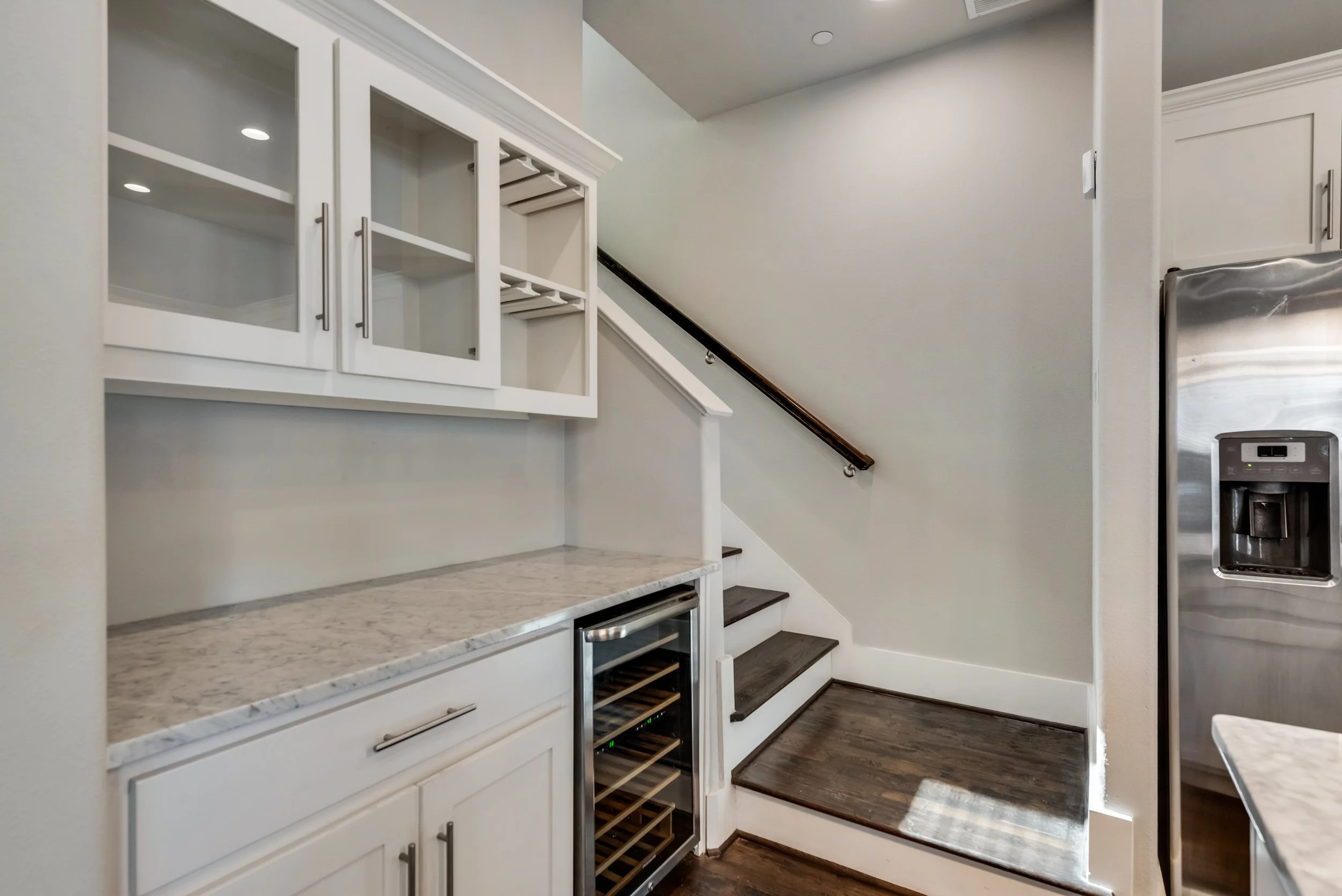 Kitchen with white cabinetry, a marble countertop, dark wooden stairs, a wine cooler, and a stainless steel refrigerator.