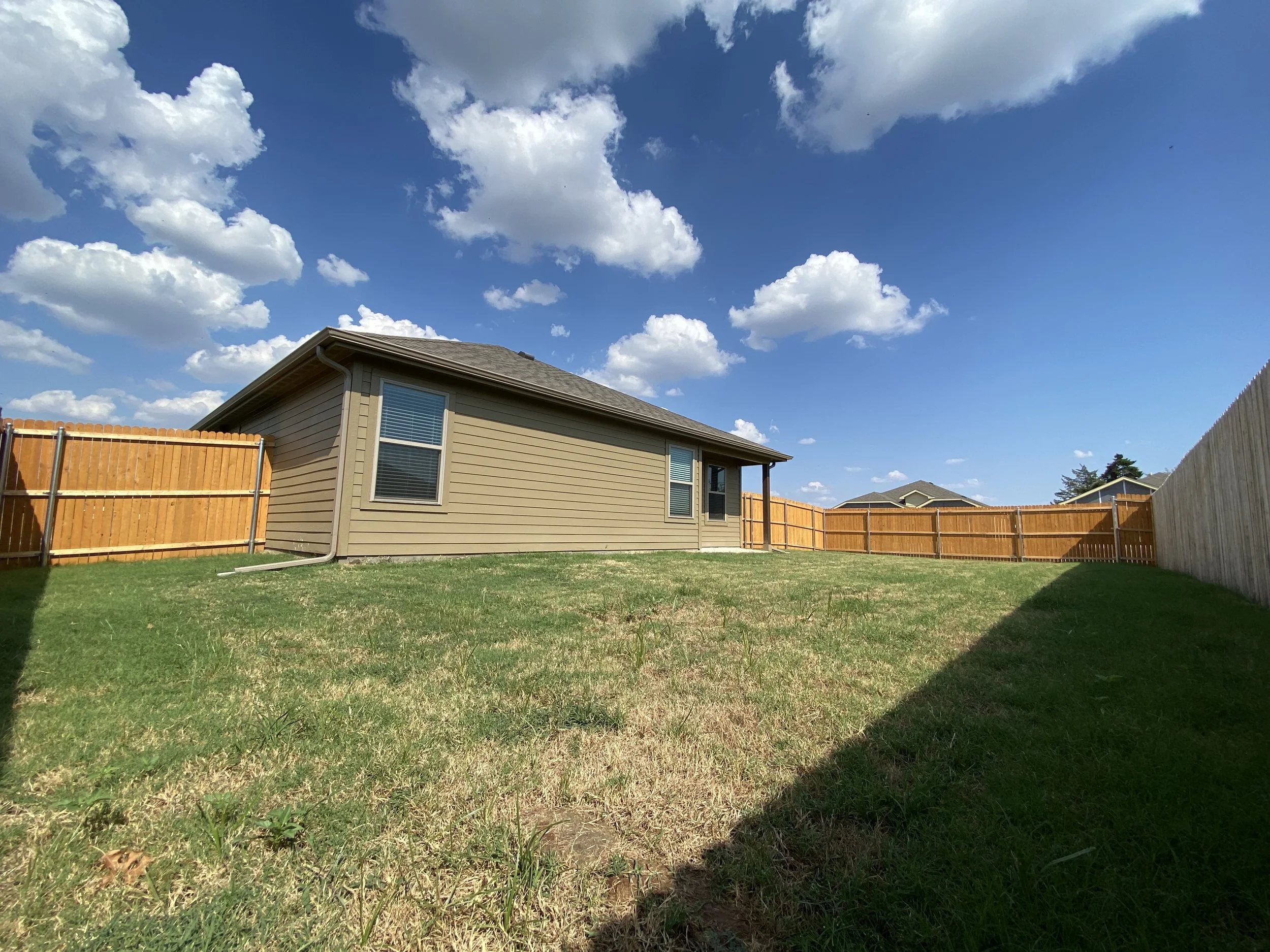 A backyard with a house, wooden fence, green grass, and blue sky with large white clouds.