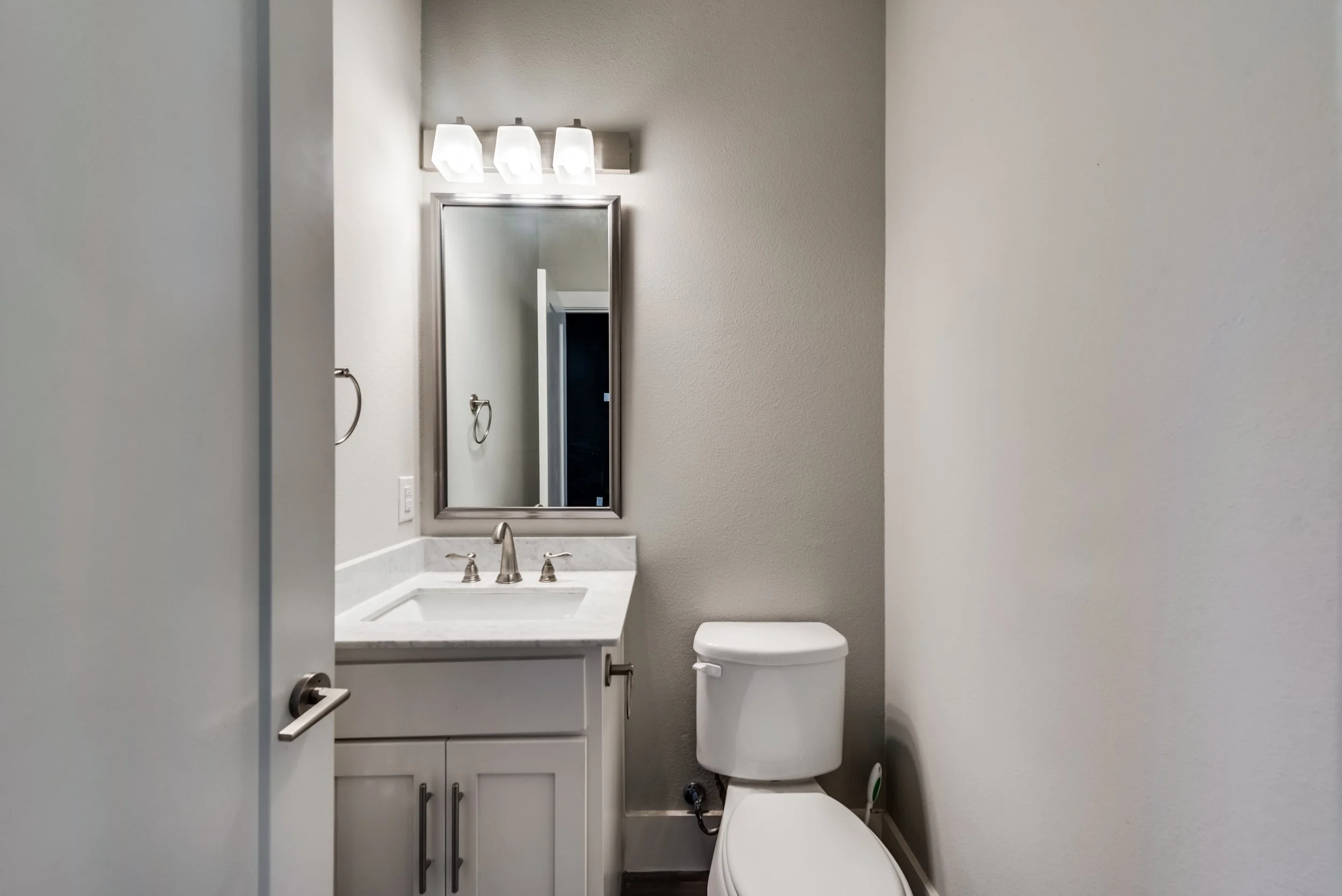 Small bathroom with a white vanity, mirror, and toilet. Light fixture with three bulbs above the mirror, cream-colored walls, and a partially visible door.
