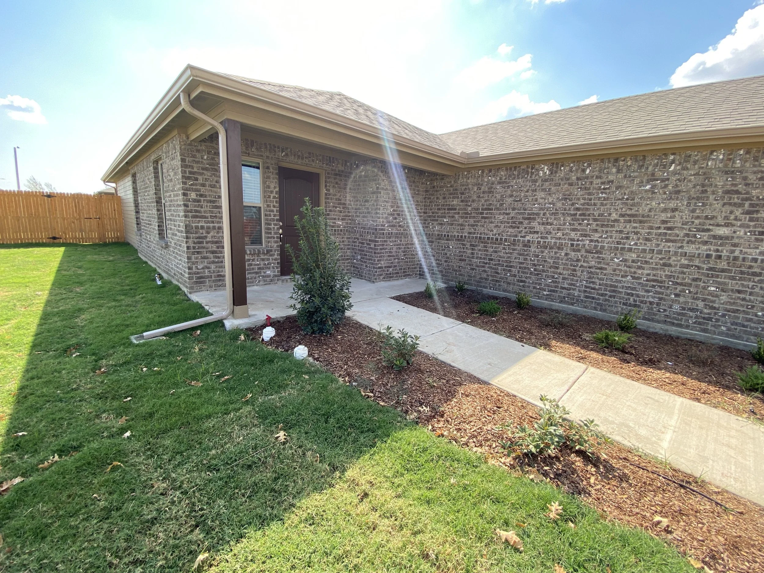 A brick house with a black door, windows, a concrete walkway, and a lawn with small bushes and plants under a partly cloudy sky.