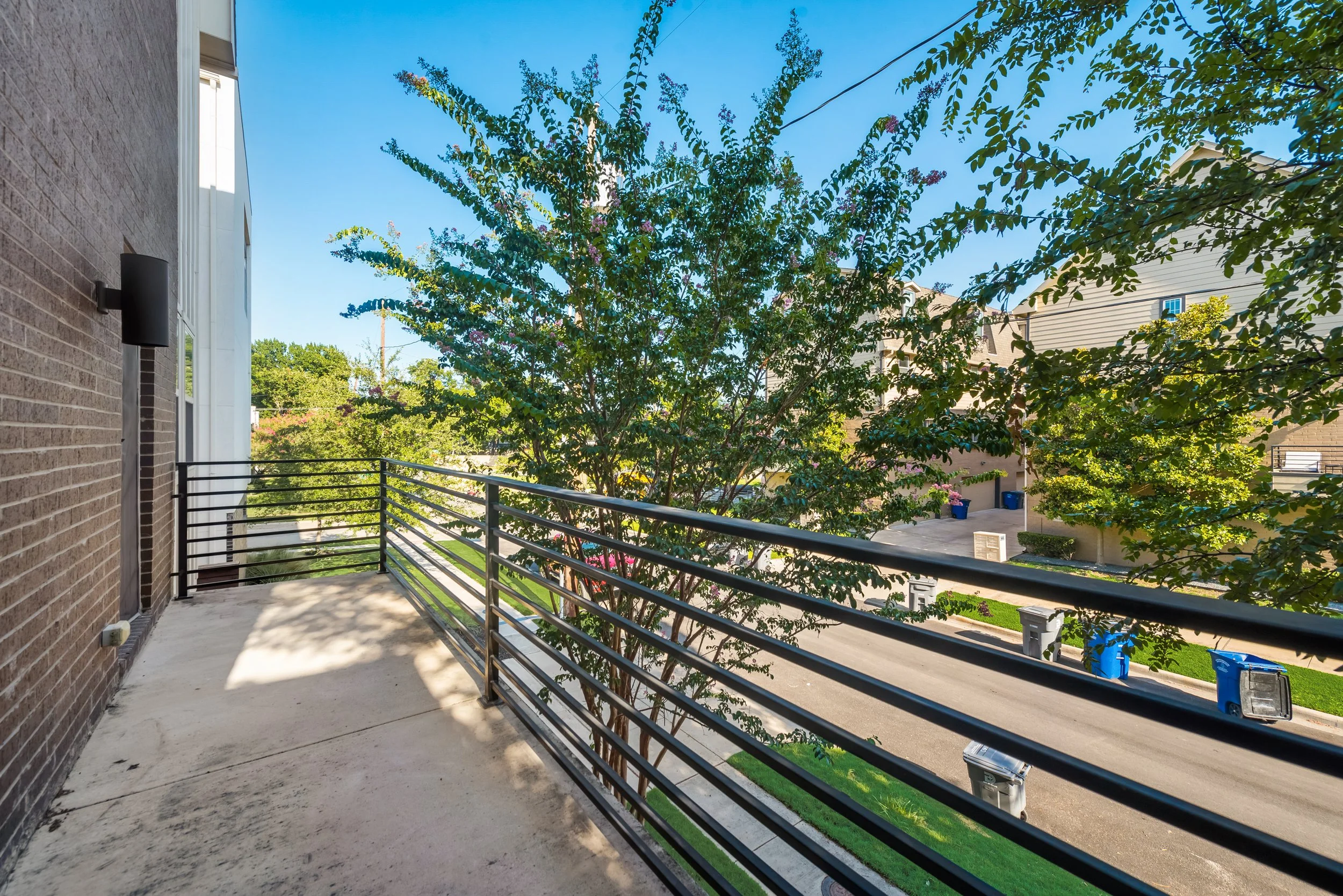 Balcony with a metal railing, tree with green leaves, and neighboring houses under a blue sky.