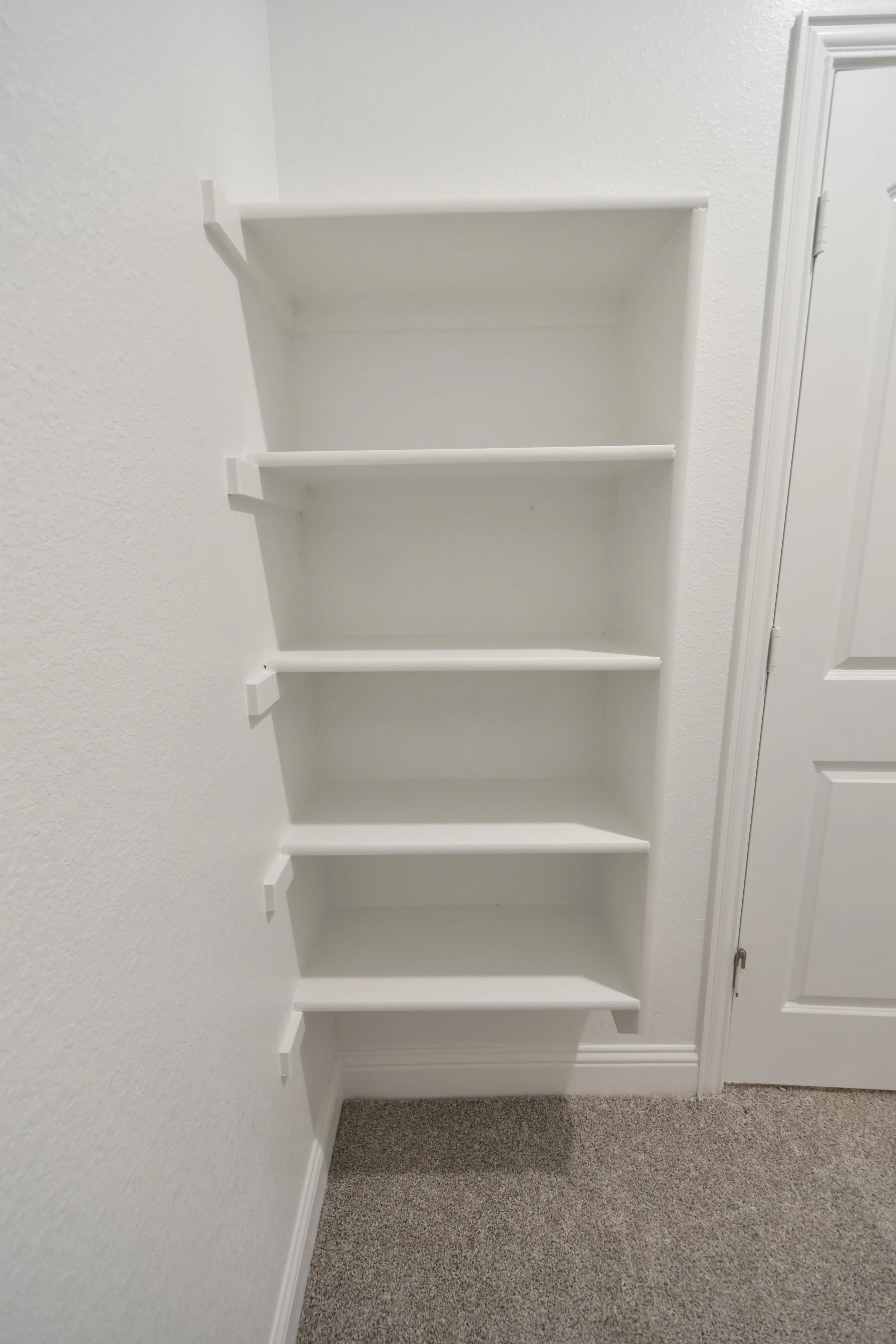 A white empty wall-mounted bookshelf with five shelves, against a white textured wall with a door on the right, with a beige carpeted floor.