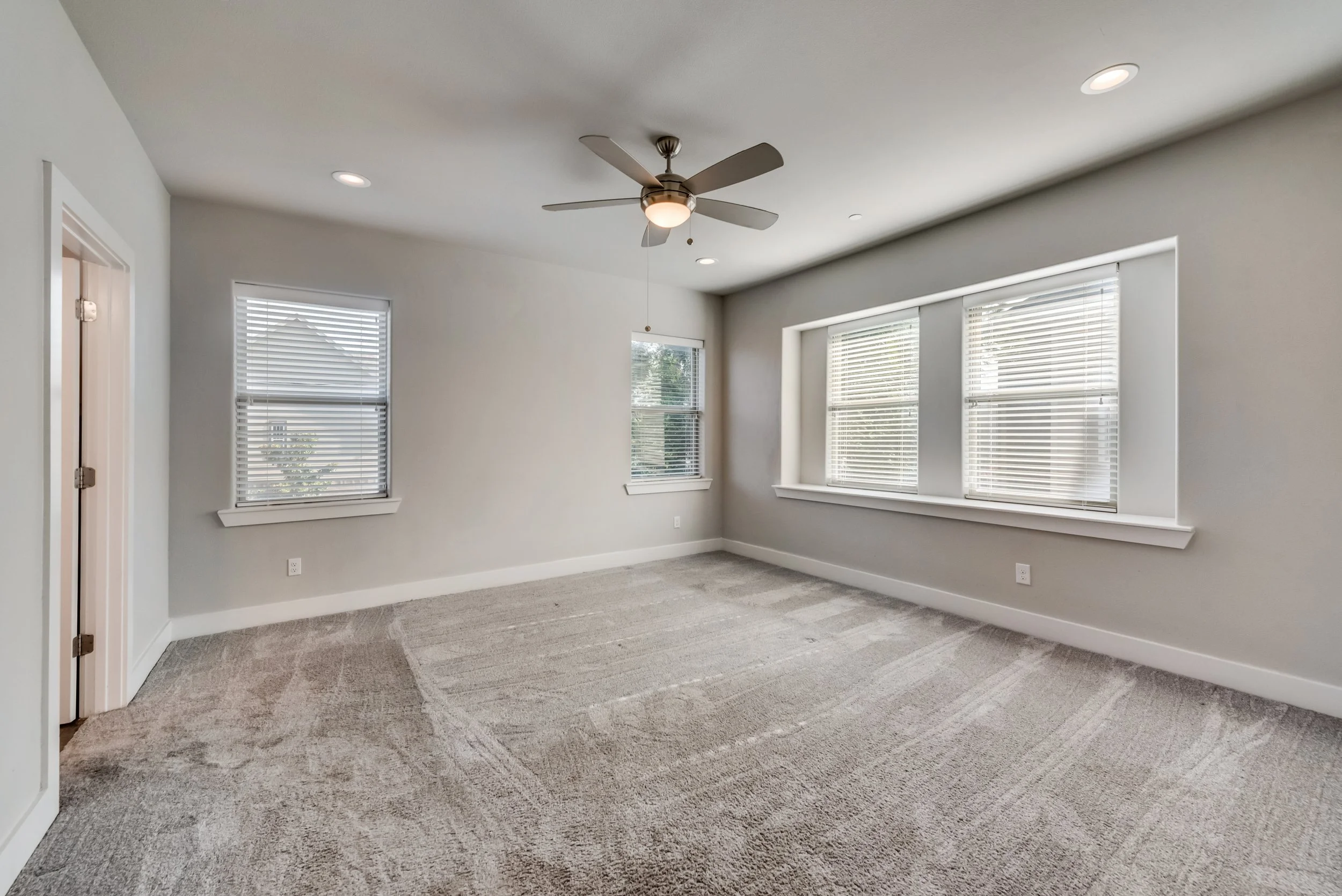 Empty room with large windows, grey carpet, white walls, ceiling fan, and recessed lighting.