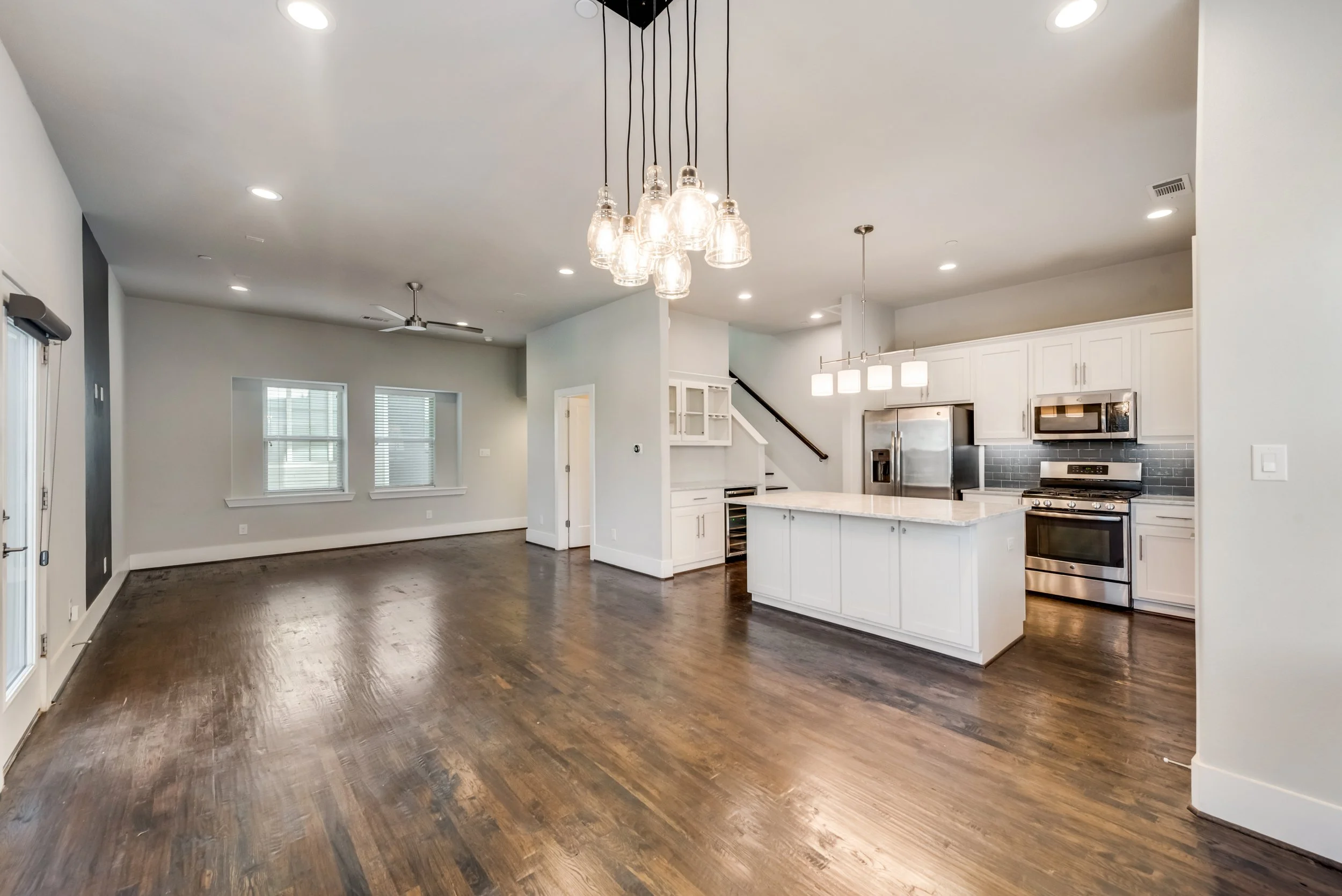 Open-concept living room and kitchen with white cabinets, stainless steel appliances, hardwood floors, large windows, modern pendant lighting, and a ceiling fan.