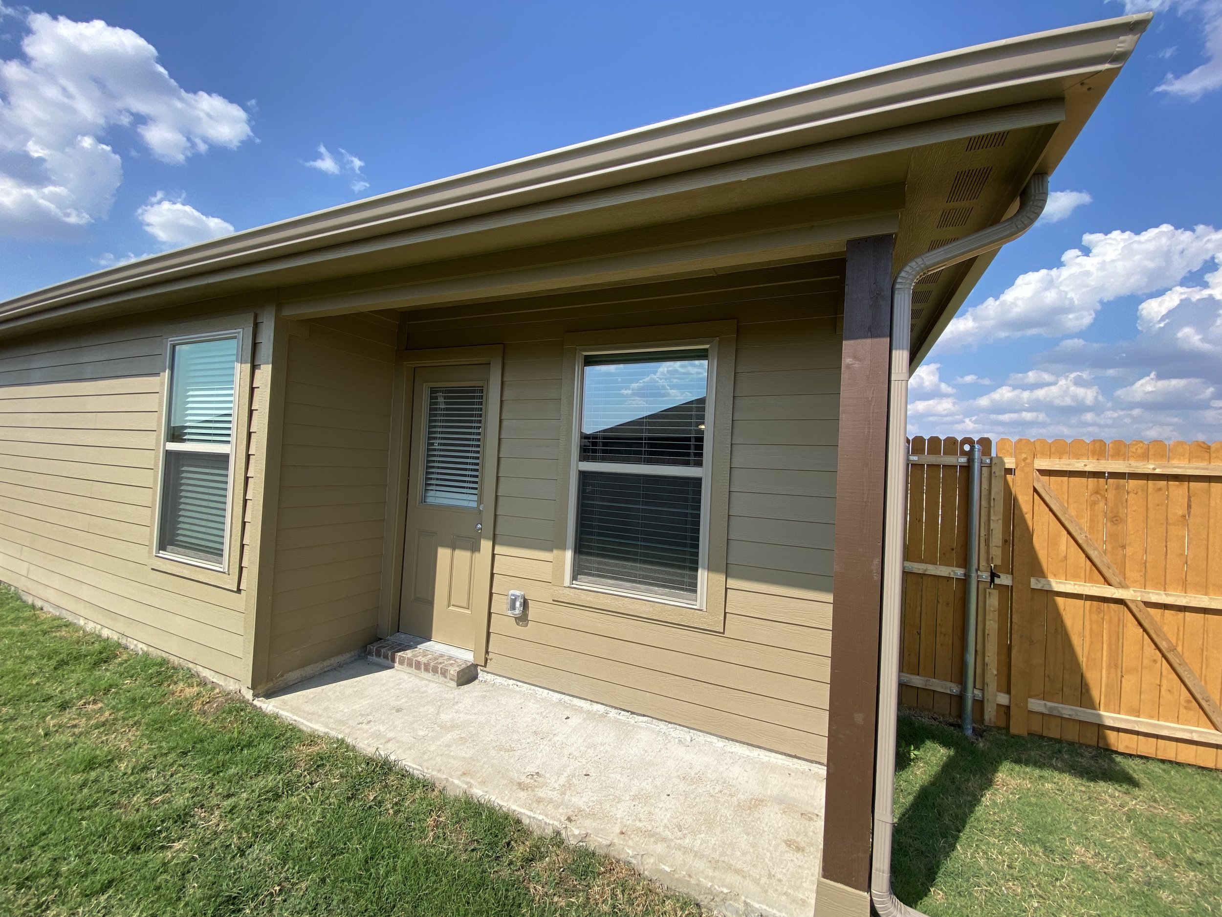 The back of a house with beige siding, a white door, and two windows. There is a concrete patio, a wooden fence on the right, and a blue sky with clouds overhead.