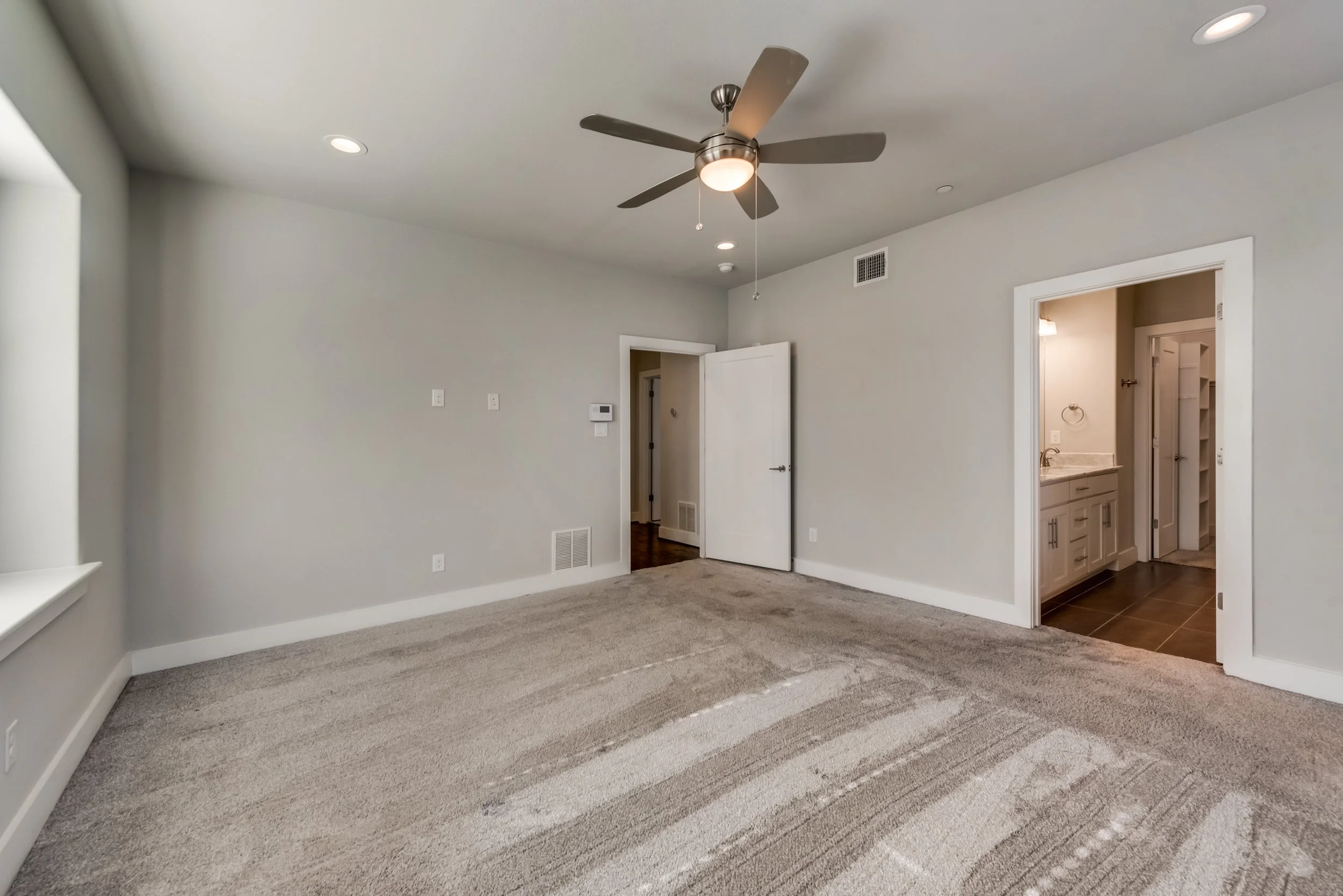 Interior of a modern, empty living room with beige carpet, light gray walls, a ceiling fan, and a doorway leading to a bathroom with white vanity and dark flooring.