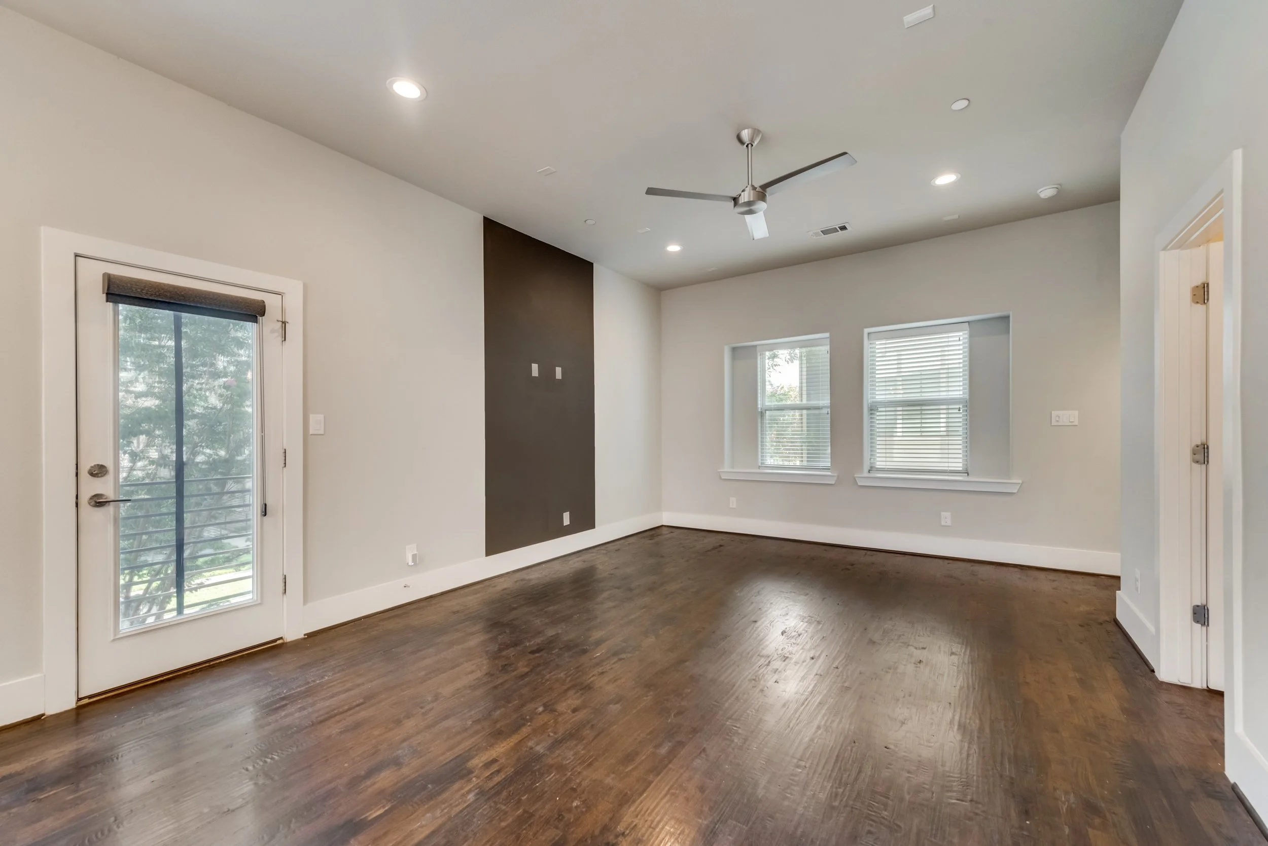 Empty room with white walls, hardwood floors, large windows with blinds, a sliding door with a screen, and ceiling lights.