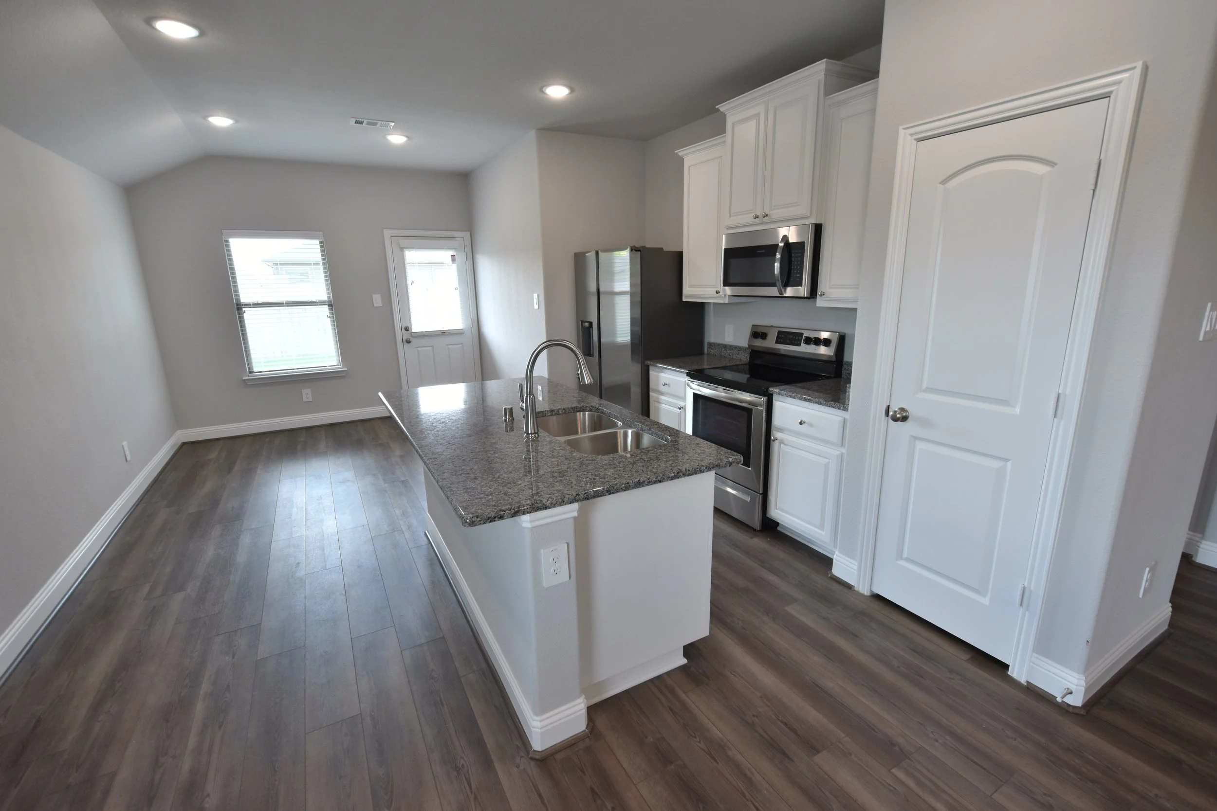 Modern kitchen with white cabinets, stainless steel appliances, a granite countertop island with a double sink, and wood flooring, with a window and a door in the background.