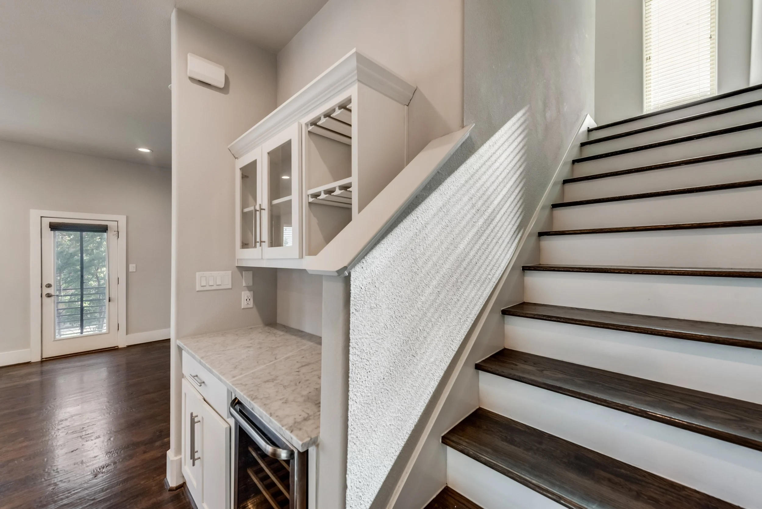 Interior of a modern home with stairs leading to an upper level, a partial view of a kitchen counter with a wine cooler, a white cabinet with glass doors, a door with a window, and hardwood floors.