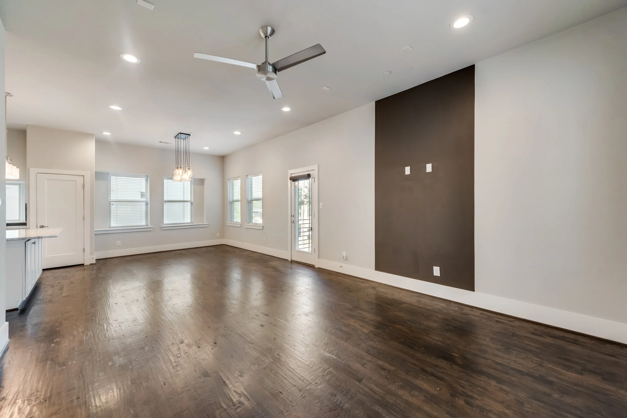 Interior of a modern, empty living room with hardwood flooring, white walls, large windows with blinds, a dark accent wall with electrical outlets, and a ceiling fan.