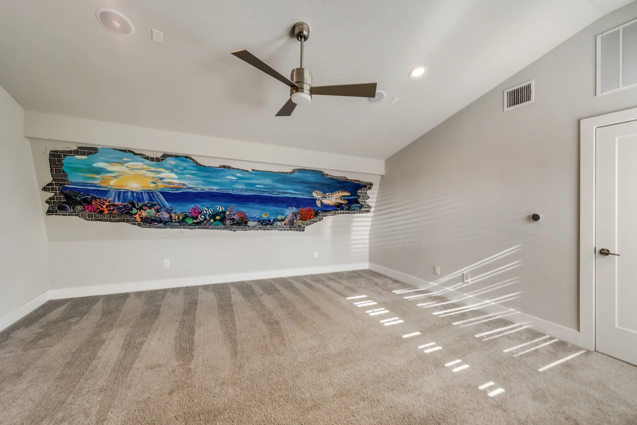 Empty room with beige carpet, white walls, ceiling fan with three blades, and a ceiling mural of an ocean scene with a sunset, seashells, and coral. Sunlight filters through window blinds, casting shadows on the floor.