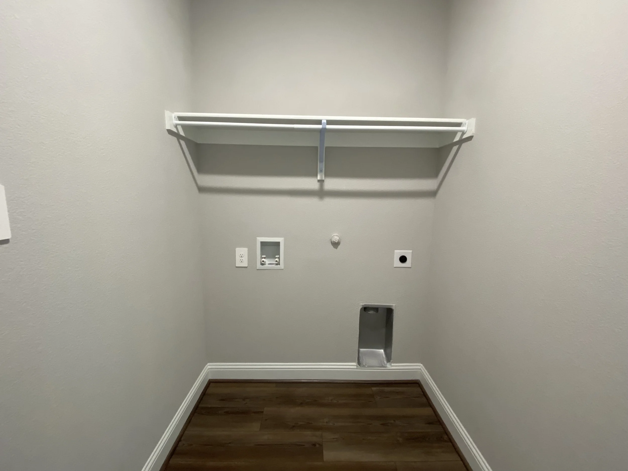 An empty laundry or utility room with light-colored walls, a white wire shelf on the wall, and outlets and plumbing for a washer and dryer, along with a small vent or dryer duct opening on the wall.