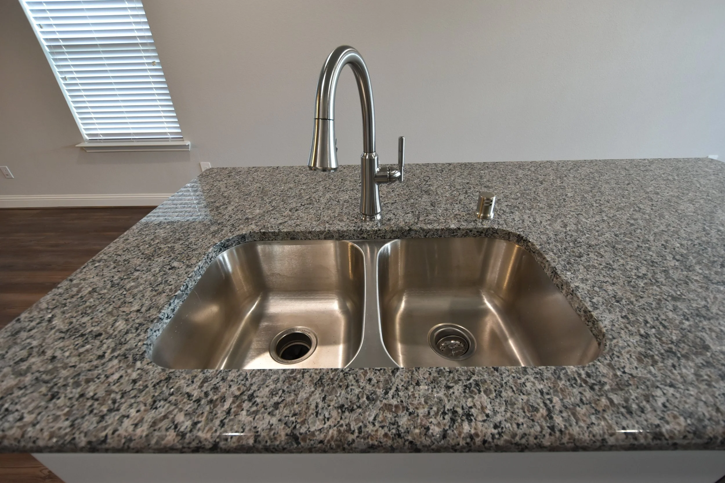 A stainless steel kitchen sink with a curved faucet and a soap dispenser on a speckled granite countertop in a kitchen.