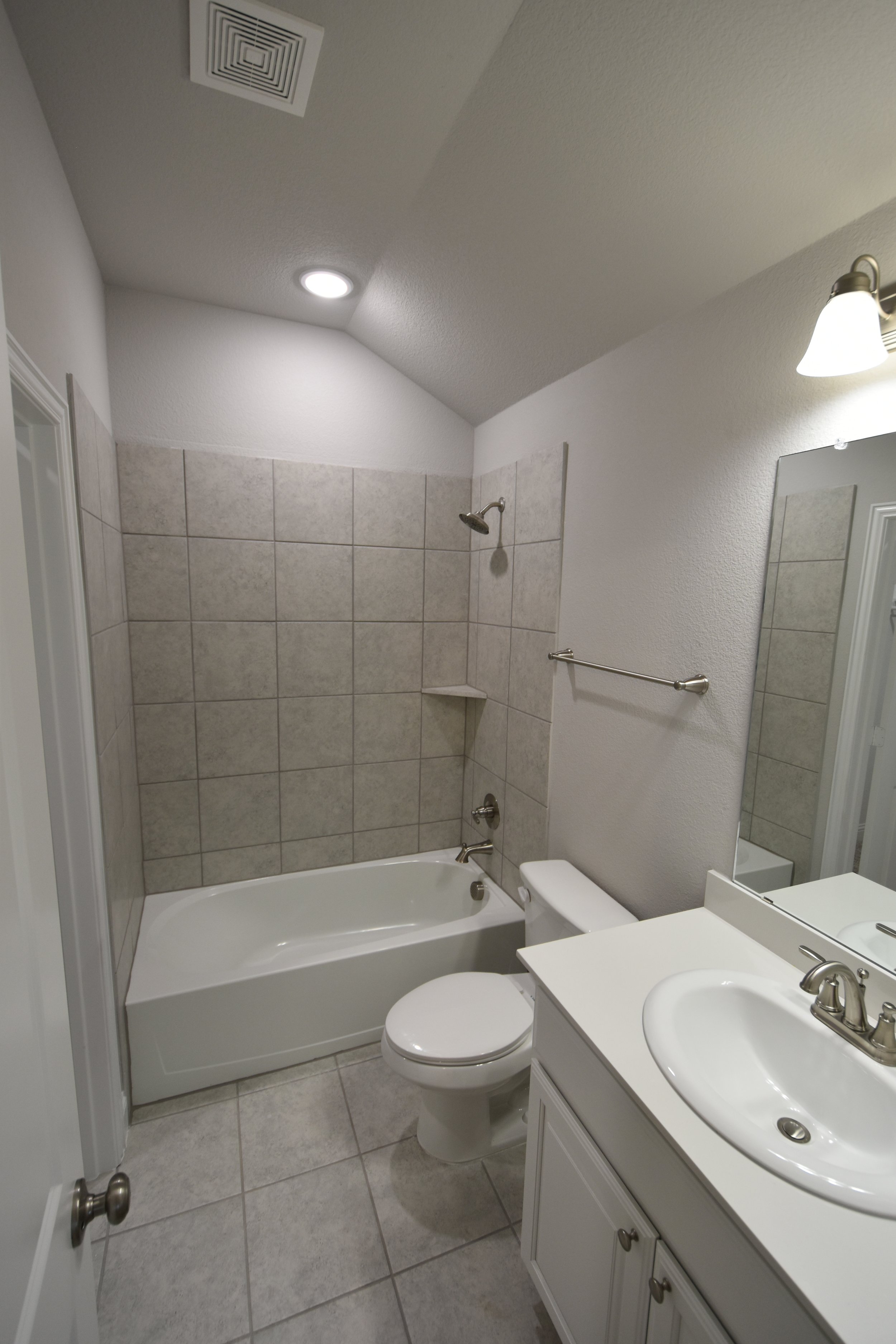 A bathroom with a bathtub, a showerhead, a toilet, a white bathroom vanity with a sink, a mirror, a towel rack, and tiled walls and floor.