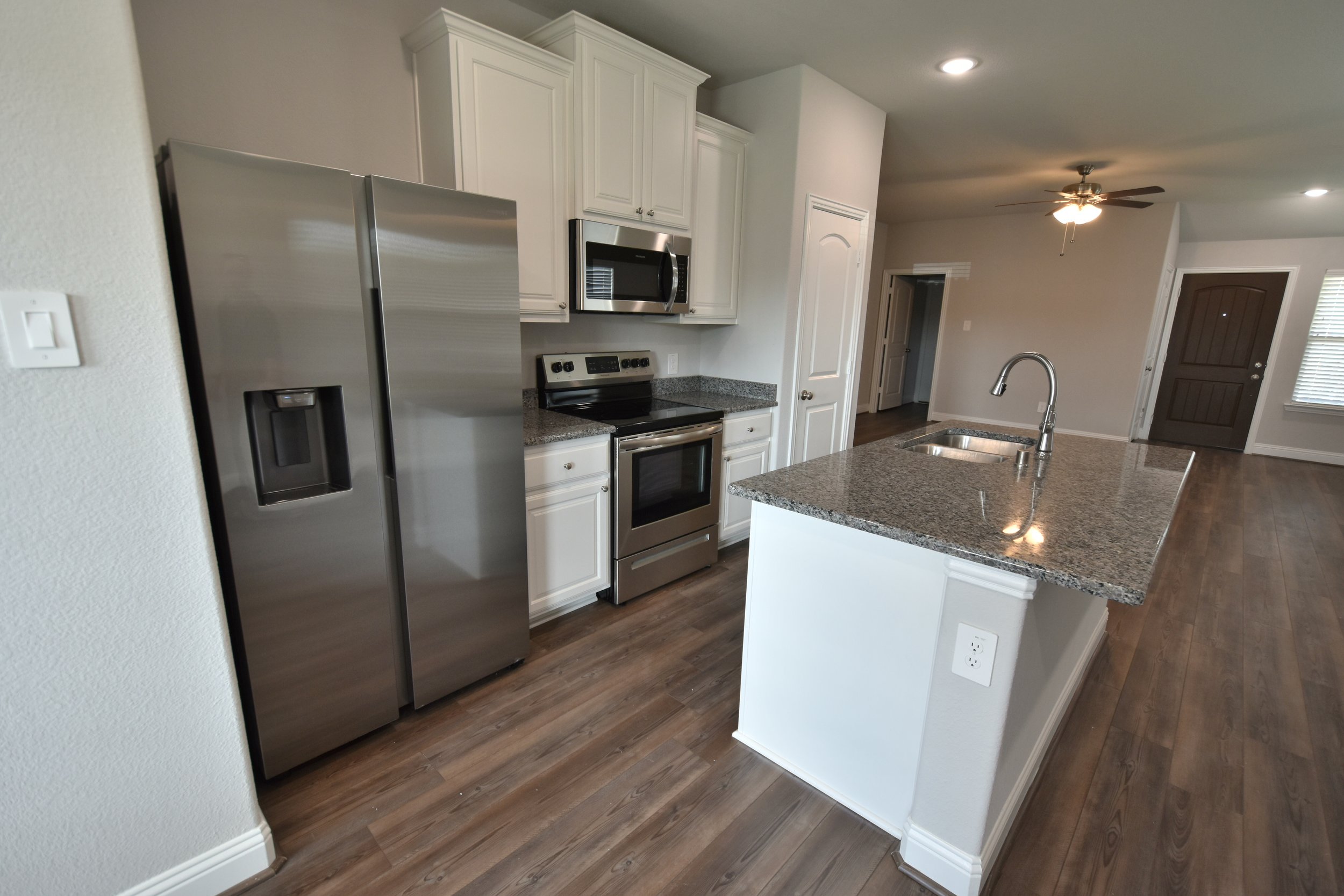 A modern kitchen with white cabinets, stainless steel refrigerator, microwave, and electric stove. Granite countertops and a central island with a sink. Hardwood floor and a ceiling fan with lights.