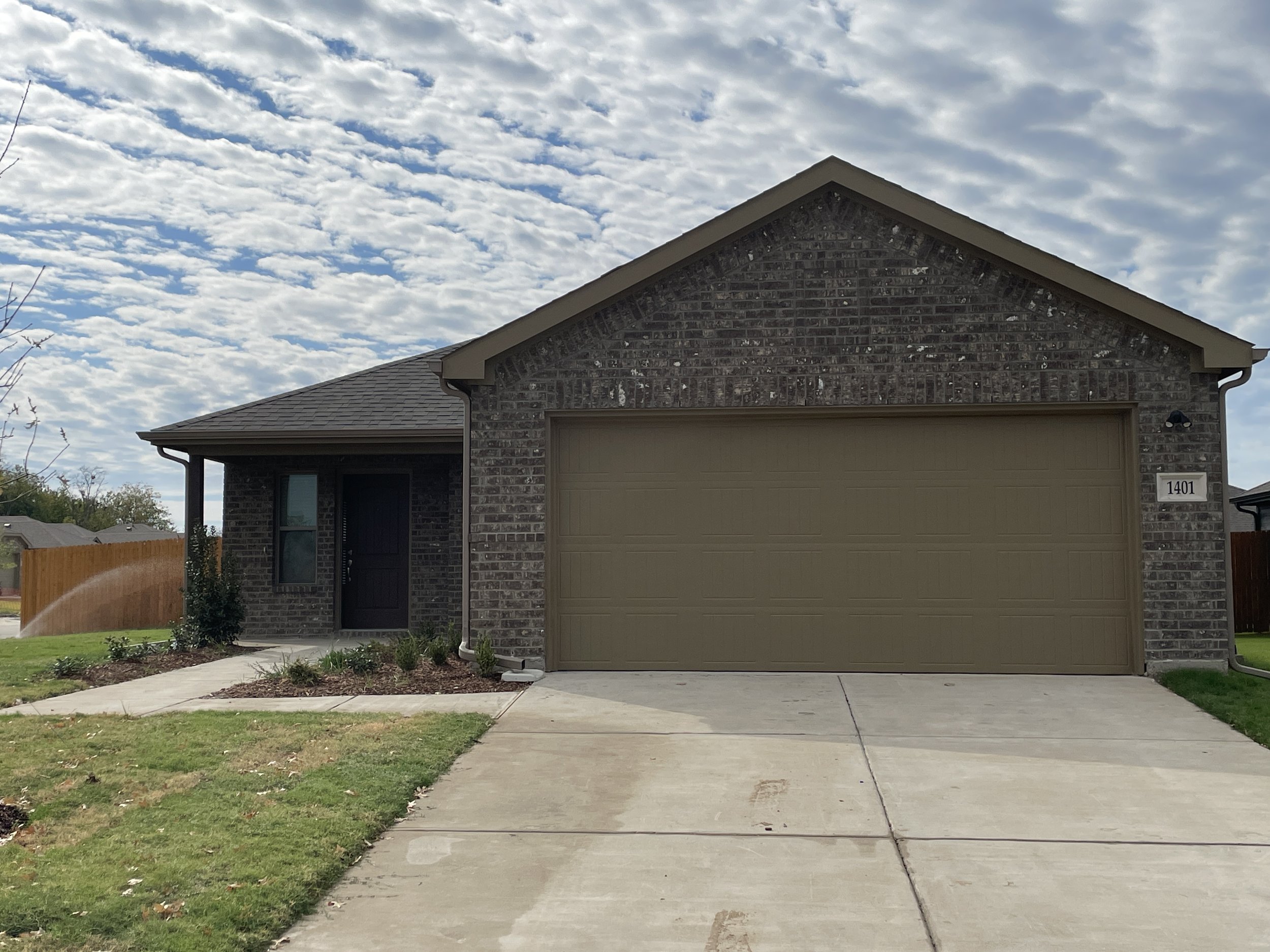 Front of a brick house with a closed beige garage door, a small porch with a black door, a window, a sidewalk and grass in the front yard, and a partly cloudy sky.