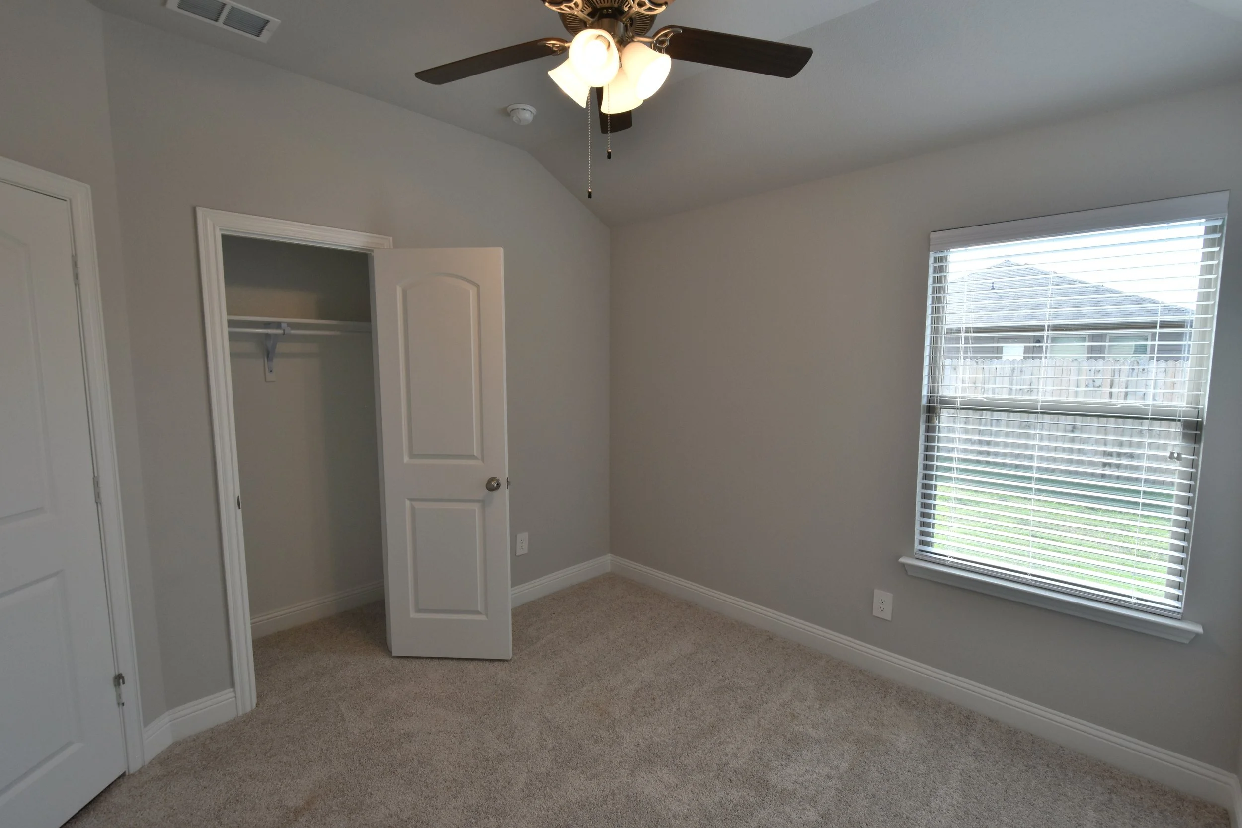 Empty bedroom with beige carpet, white walls, a window with blinds showing a fence and neighboring house outside, an open closet door, and a ceiling fan with lights.