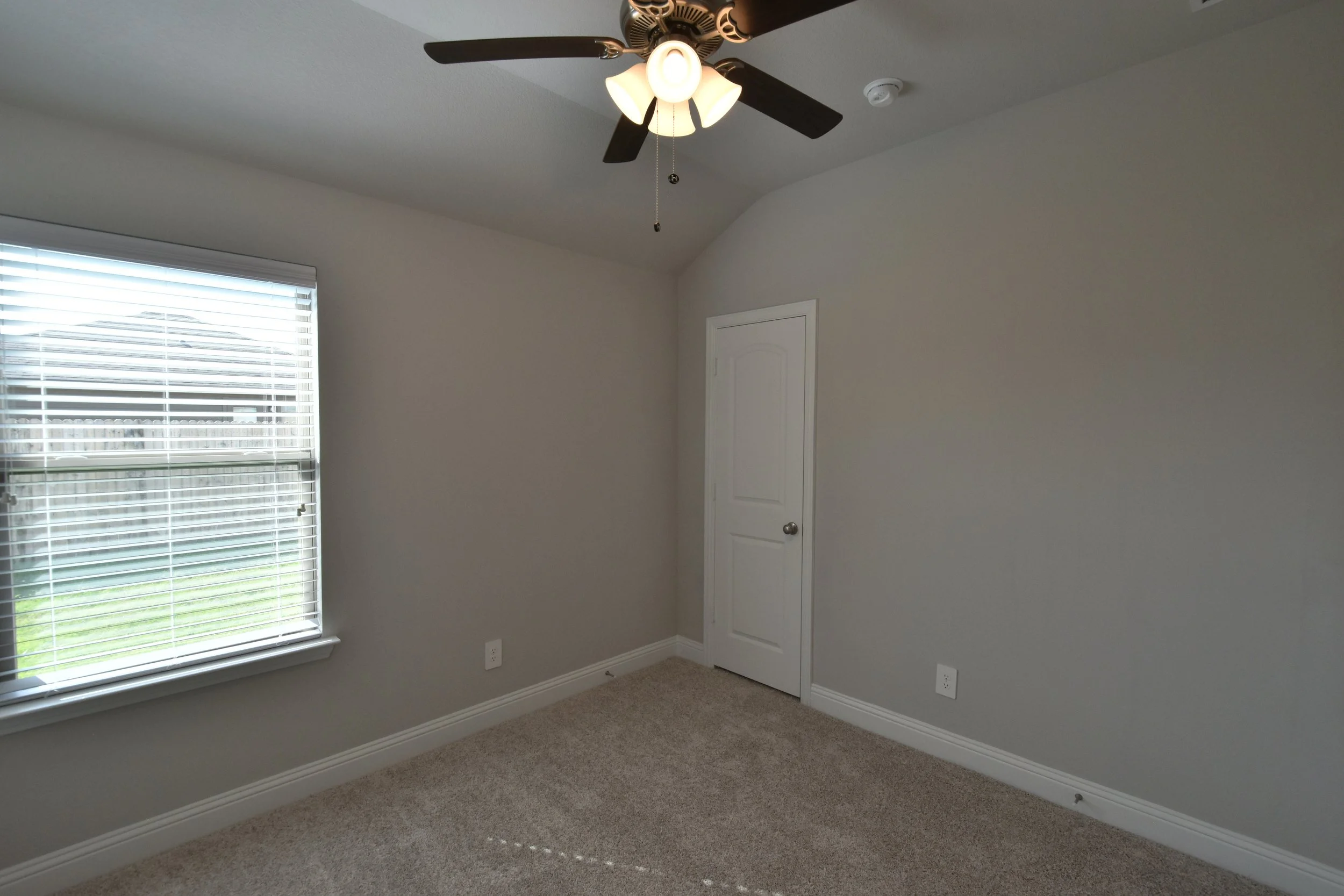 Empty room with beige carpet, light gray walls, a window with white blinds, a closed white door, and a ceiling fan with a light fixture.