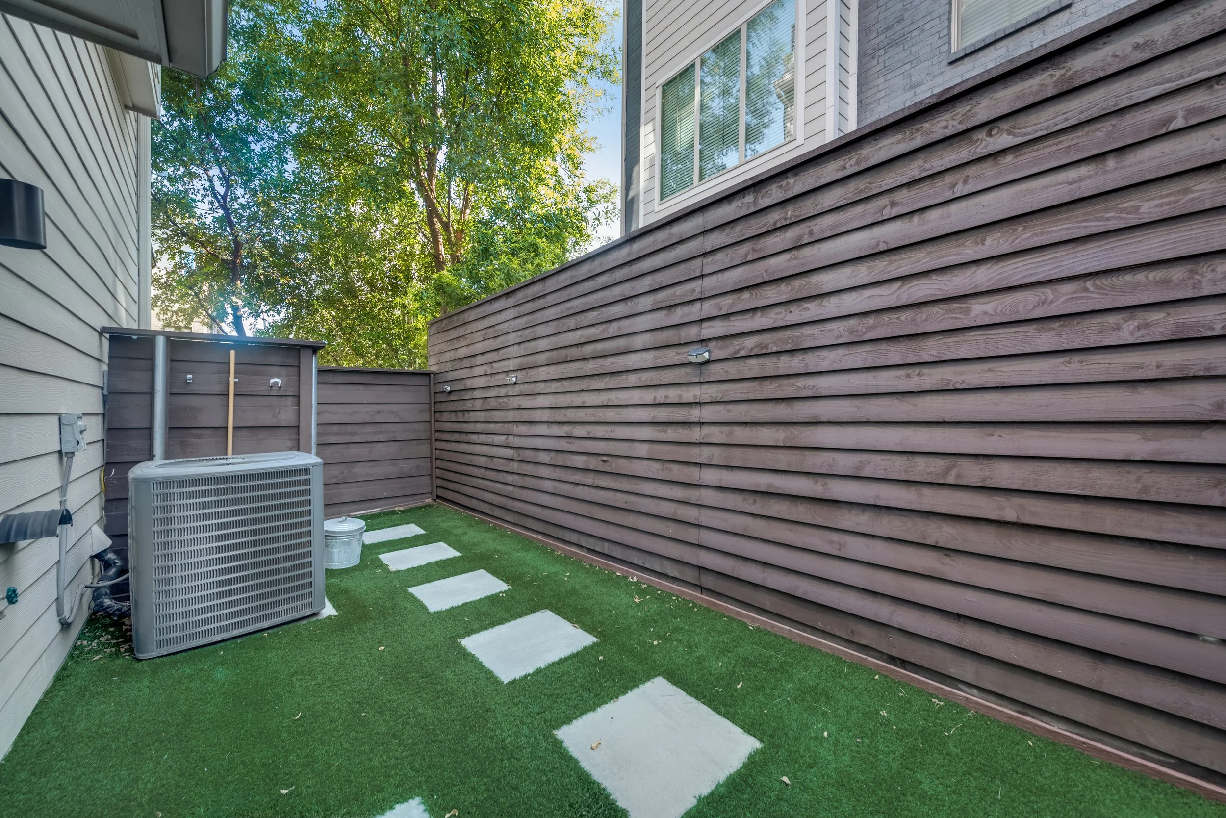 Small outdoor backyard space with artificial grass, a gray wooden fence, and part of the house with beige siding. A pathway of square stepping stones leads through the grass. There is a large tree with green leaves, and an air conditioning unit with 