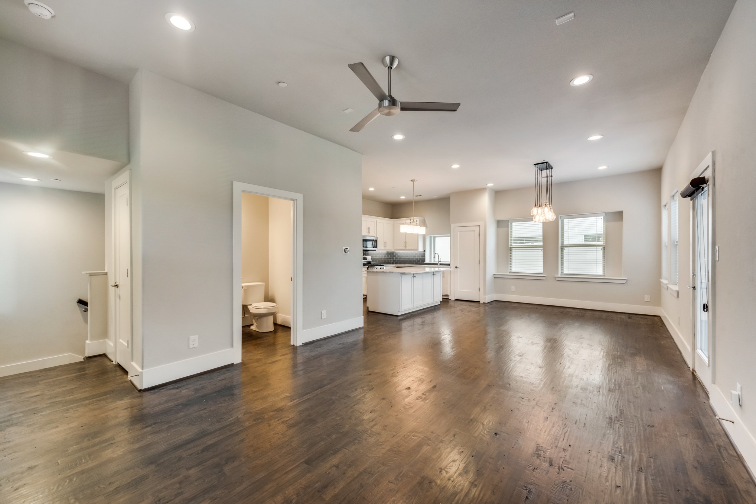 Open-concept living area with hardwood floors, white walls, ceiling fan, recessed lighting, and large windows. Kitchen with white cabinets, gray backsplash, and island.