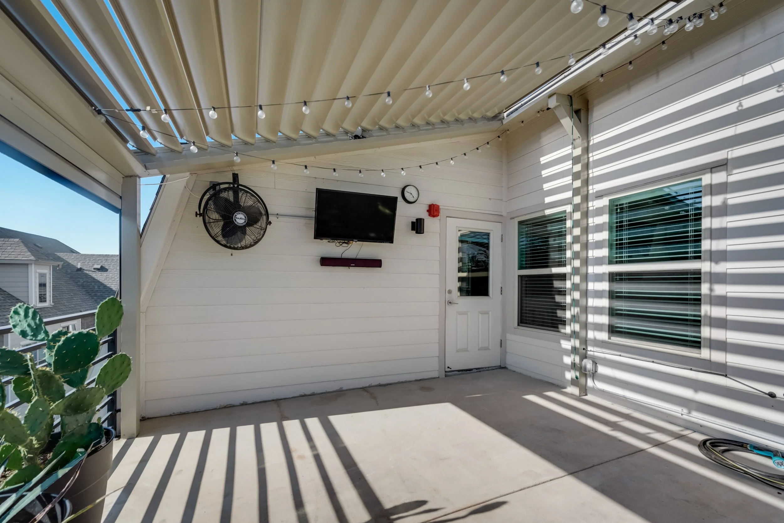 A covered outdoor patio area with white siding, string lights hanging from the ceiling, a wall-mounted television, a clock, a fan, windows with blinds, and a door, with some potted plants and shadows cast by the lights.