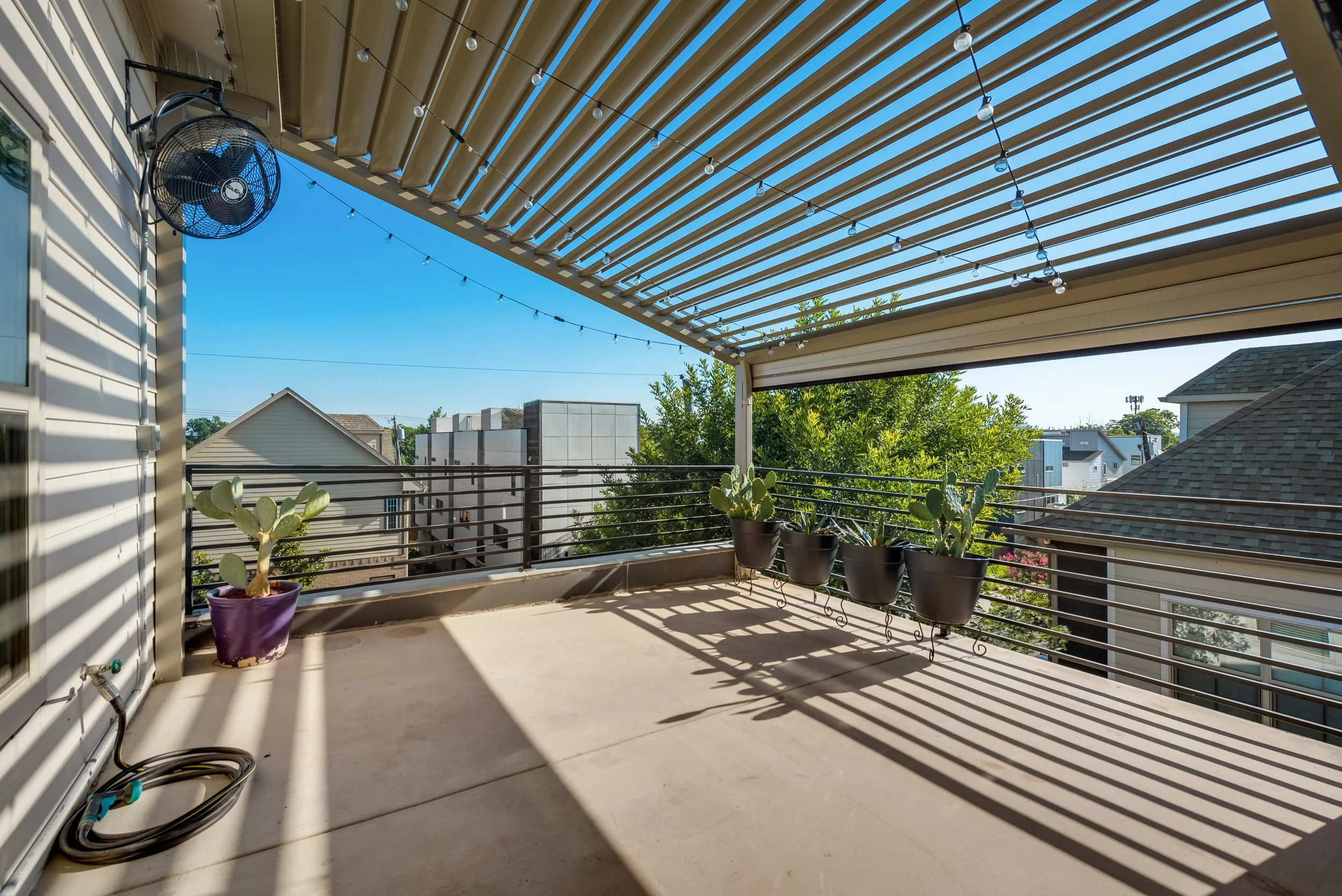 A covered balcony with string lights hanging from the ceiling, potted cacti on a metal railing, and a wind fan attached to the house wall. The sky is clear and blue with neighboring rooftops visible.