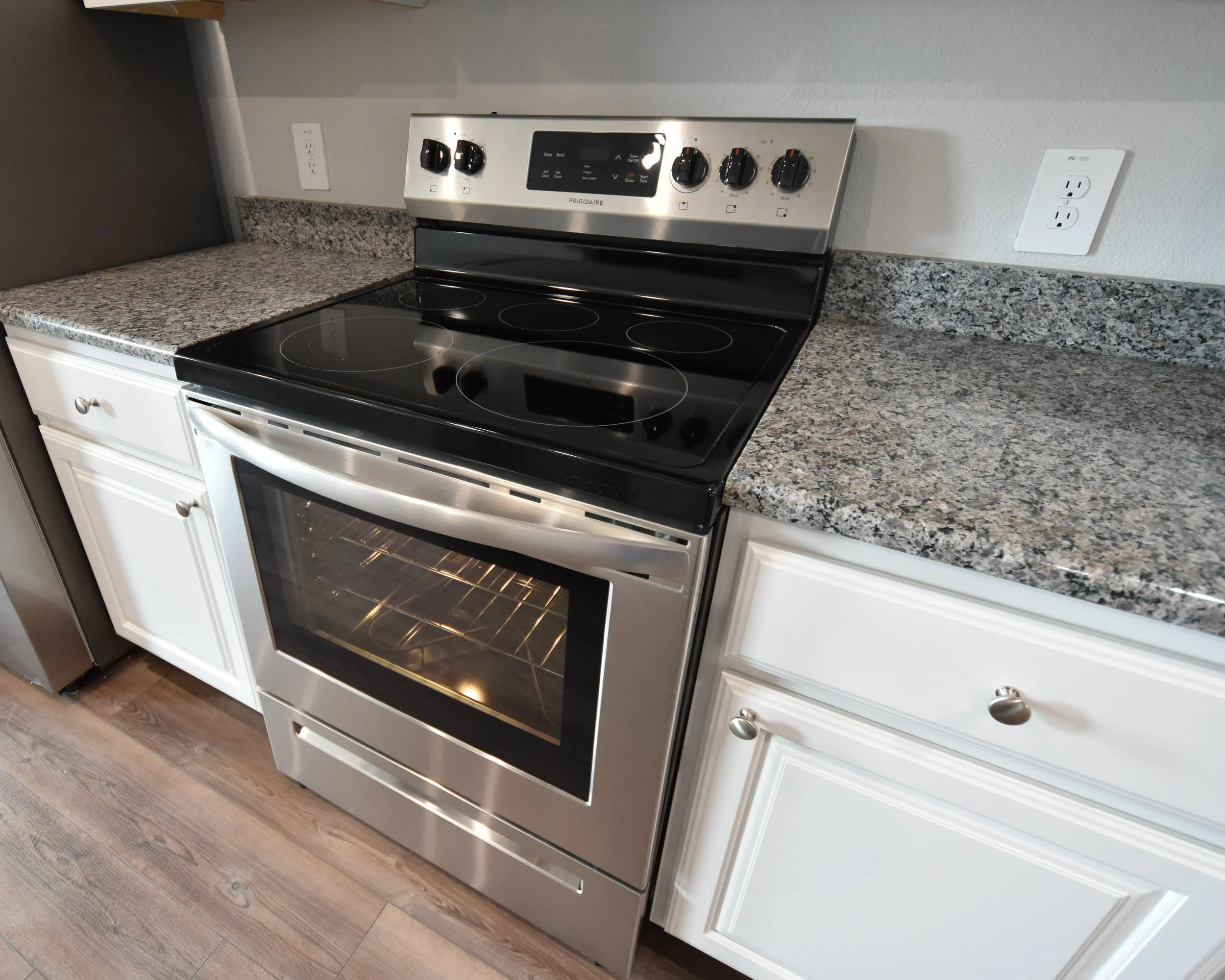 Stainless steel electric stove with black glass cooktop, digital display, and control knobs, set in a kitchen with white cabinets and granite countertops.