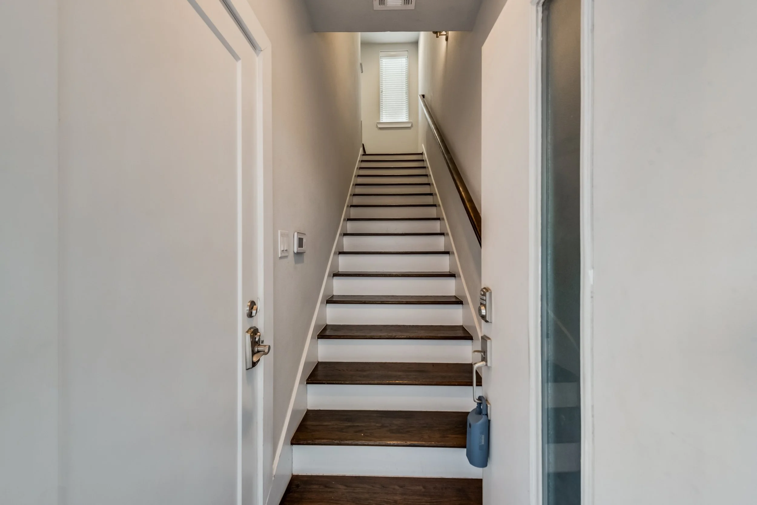 A staircase with dark wood steps, white risers, a wooden handrail on the right, and beige walls, leading up to a bright window with blinds at the top.