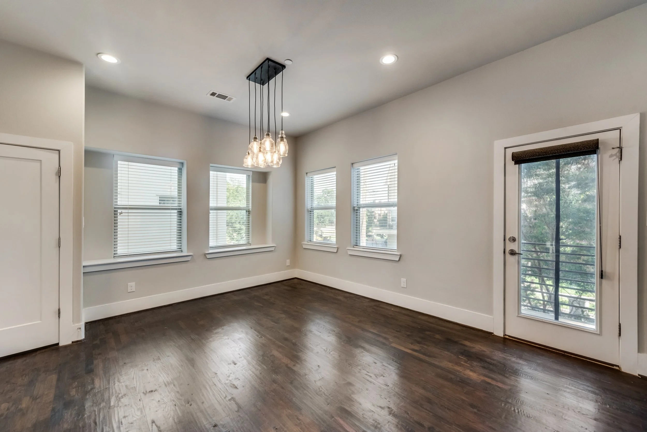 Empty living space with hardwood floors, white walls, multiple windows with blinds, a glass door, and a modern hanging light fixture.