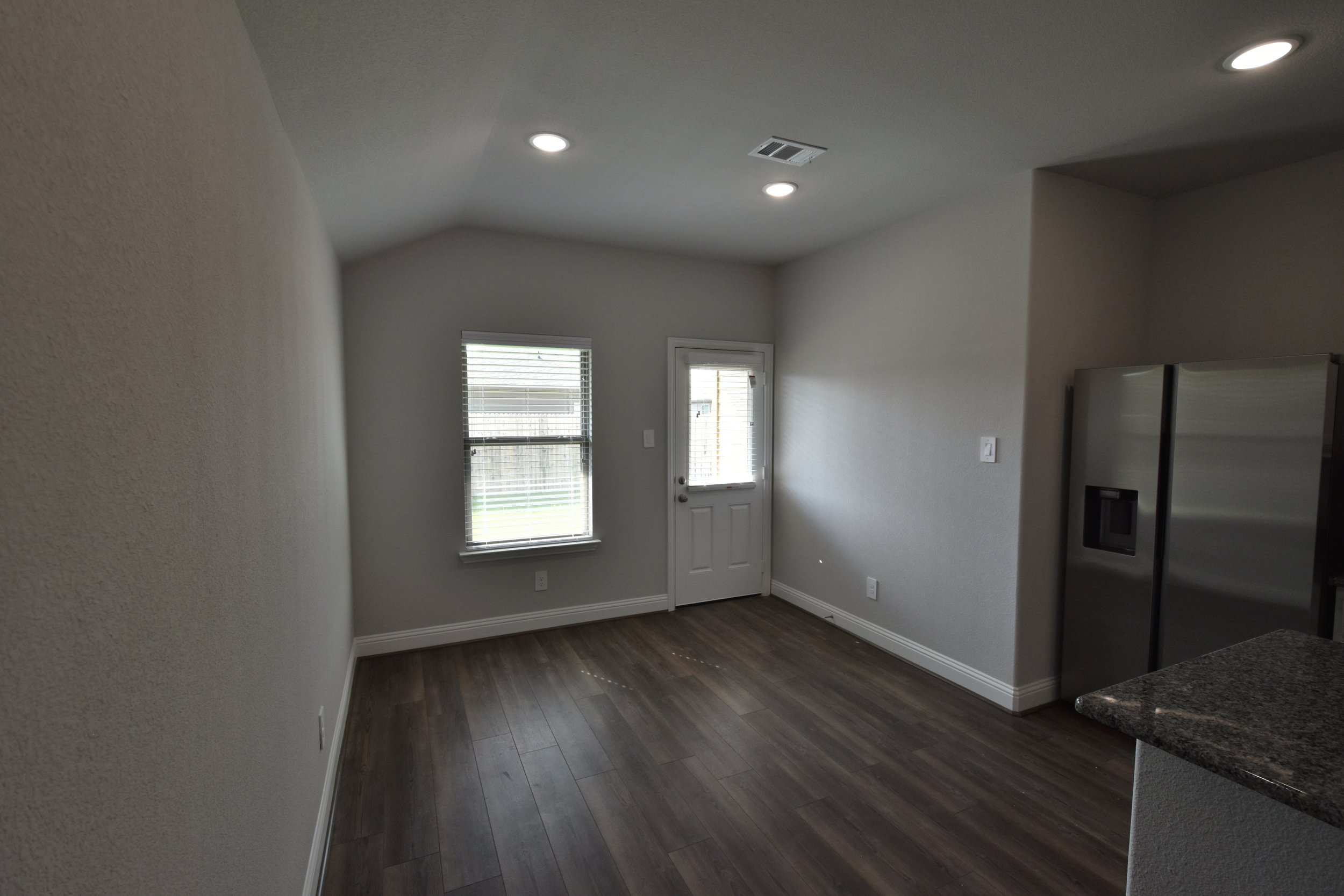 Interior of a room with a door and window, white walls, and dark wood flooring, partially visible kitchen counter with granite surface.