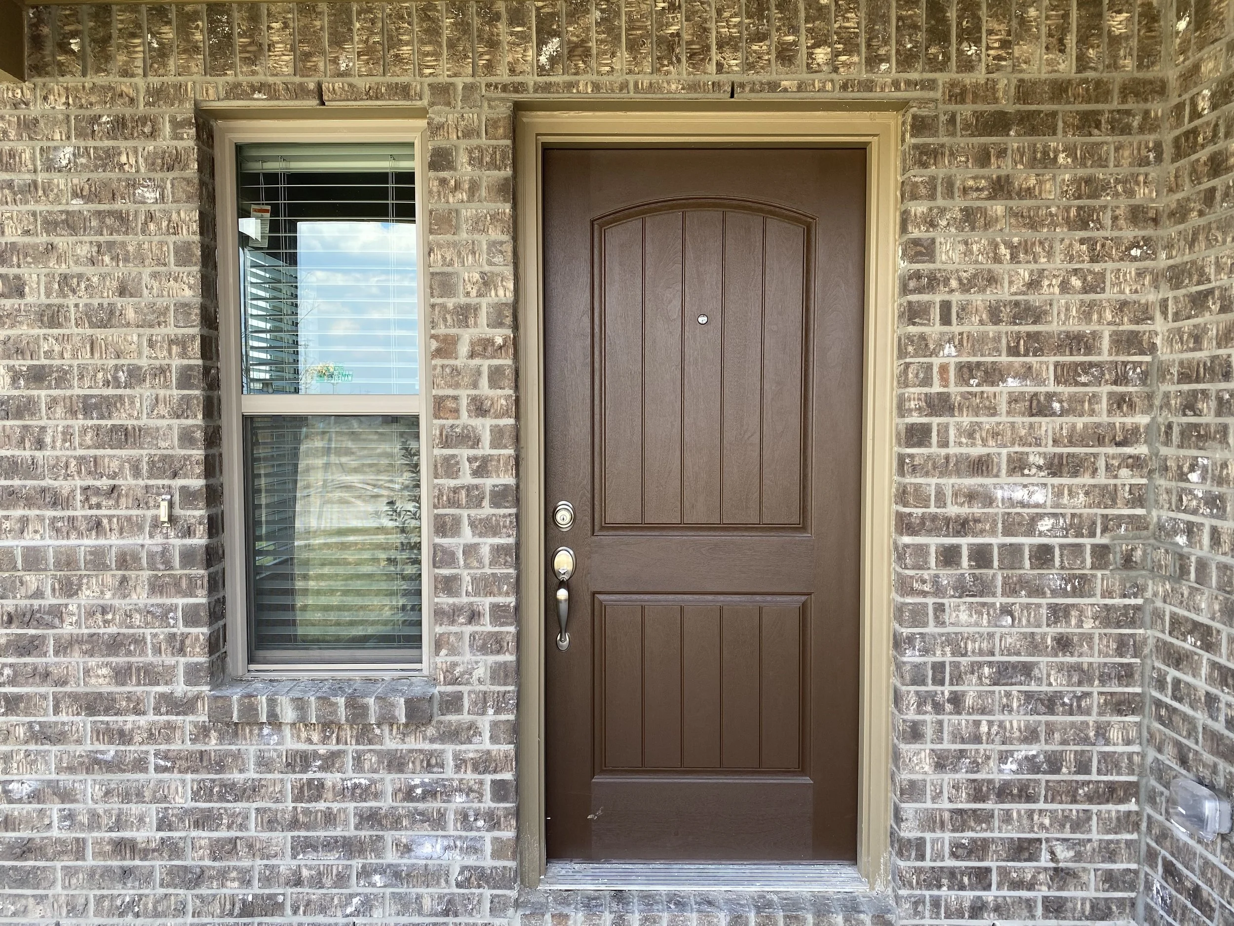 A brick house front door with a dark brown wooden door and a silver handle, surrounded by light-colored trim and a window with blinds to the left, reflecting a partly cloudy sky.
