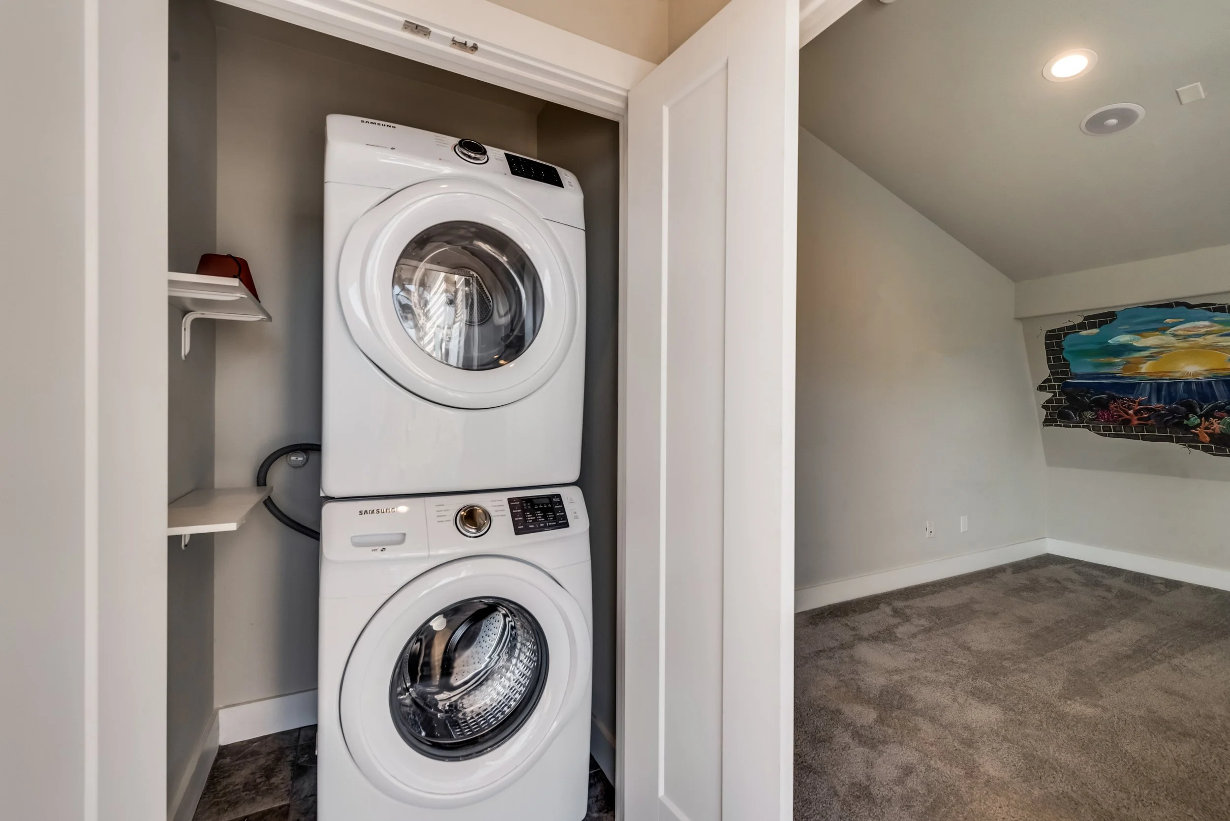 A stacked white washing machine and dryer inside a small laundry closet with open doors, adjacent to a beige wall with a partial mural of a landscape scene and a gray carpeted floor.