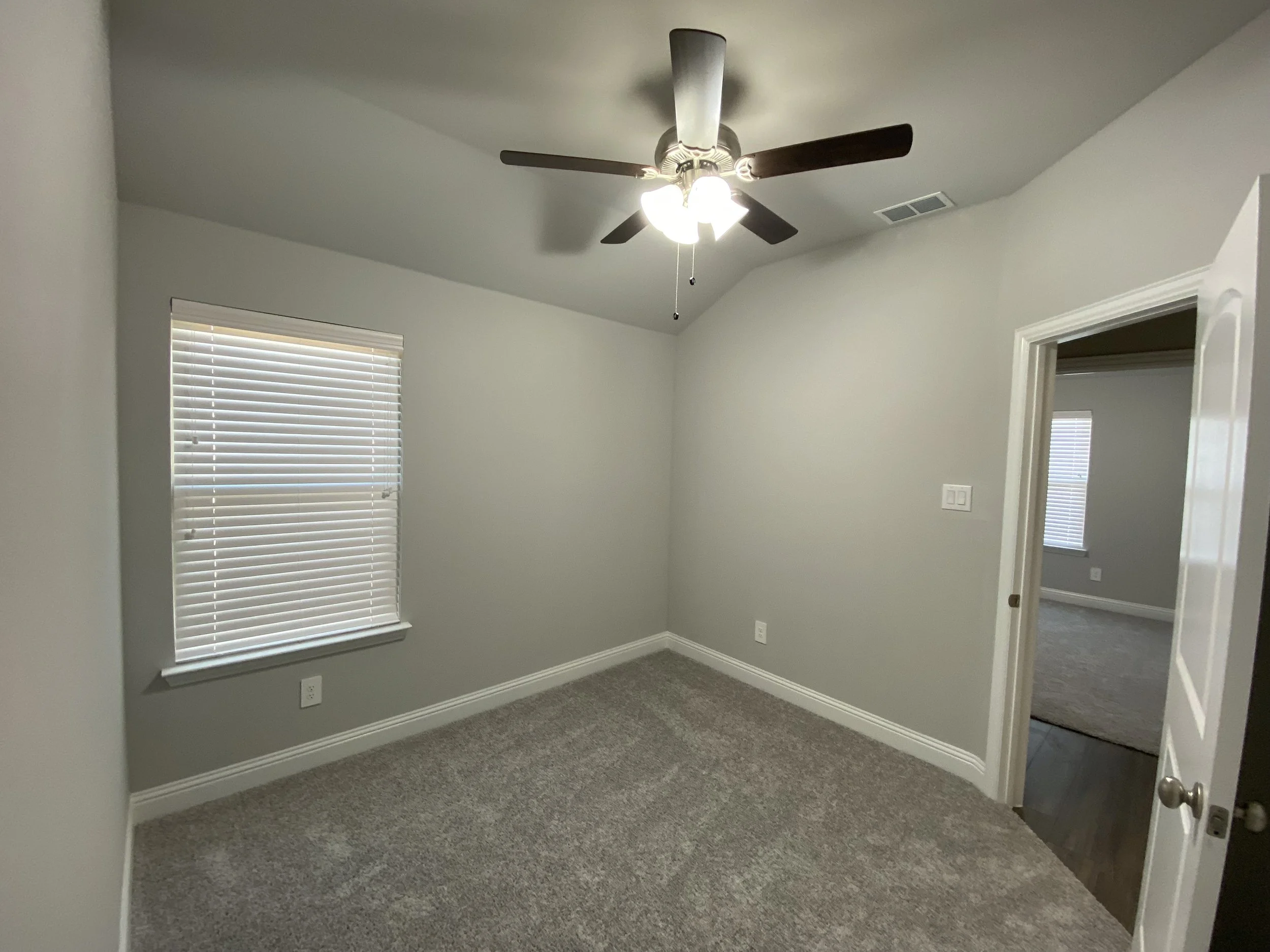 Empty room with beige carpet, gray walls, a window with blinds, a ceiling fan with lights, and an open door leading to a hallway.