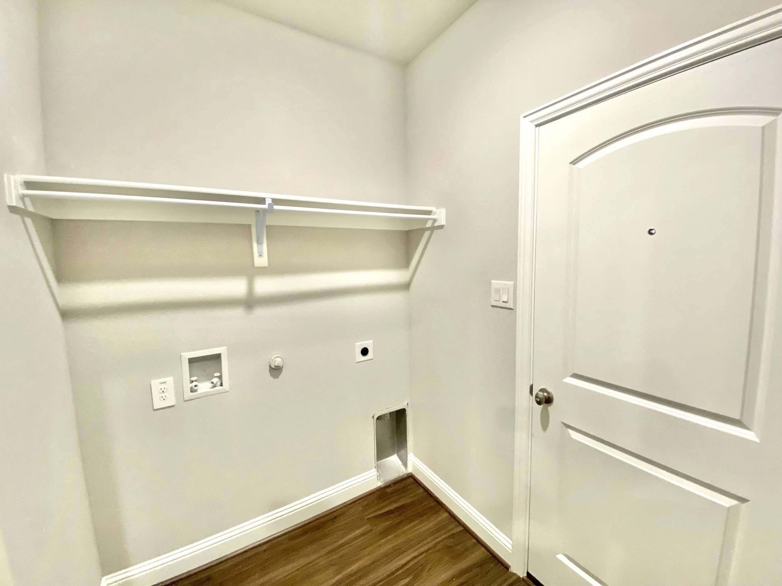 Empty laundry or utility closet with a white wire shelf mounted on the wall, electrical outlets, and a hole in the wall for plumbing or utilities, with a white door on the right.