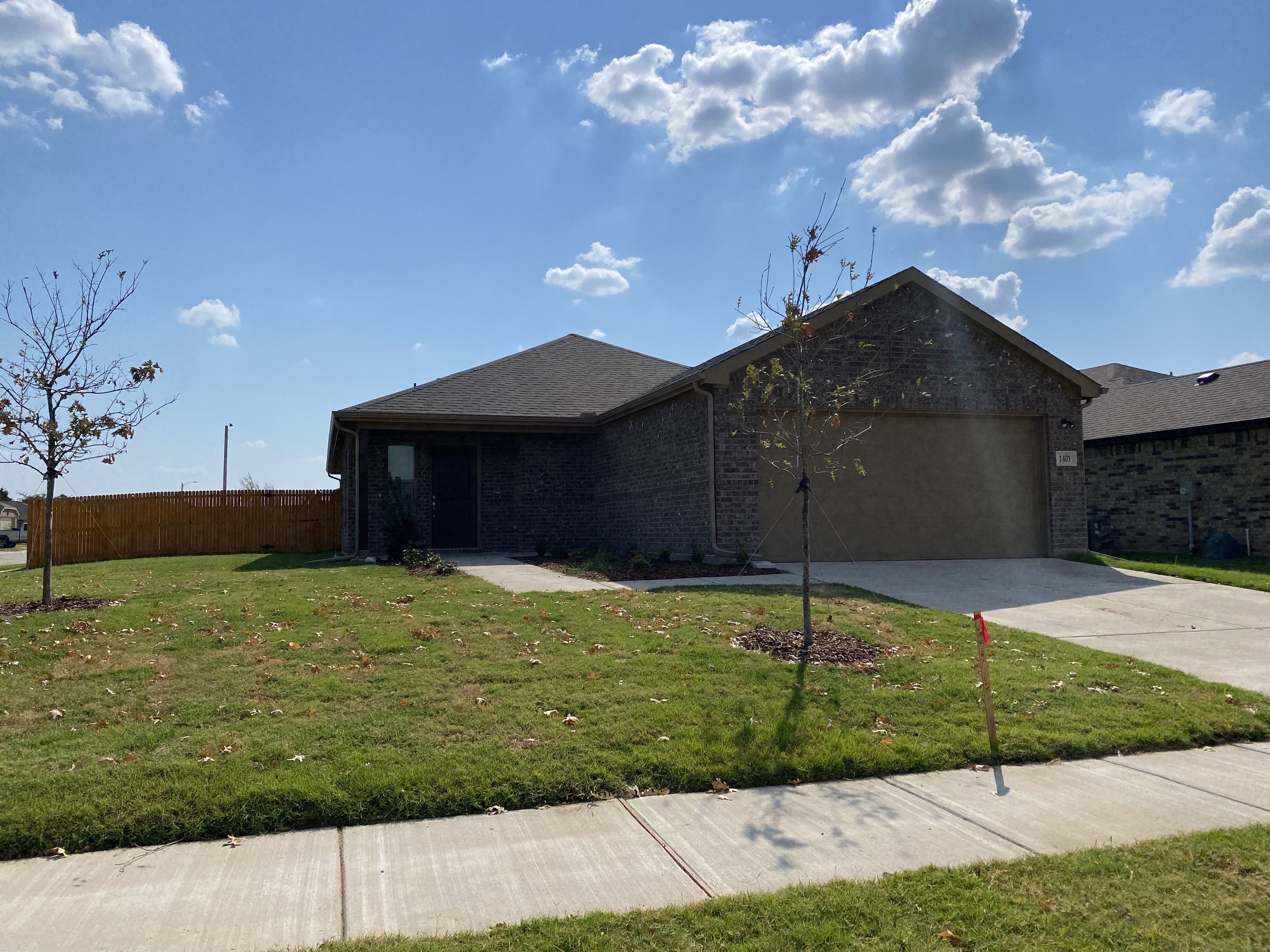 A suburban house with a brick exterior and a two-car garage, in a yard with two small trees, a wooden fence to the left, a sidewalk, and a grassy lawn. The sky is blue with scattered clouds.