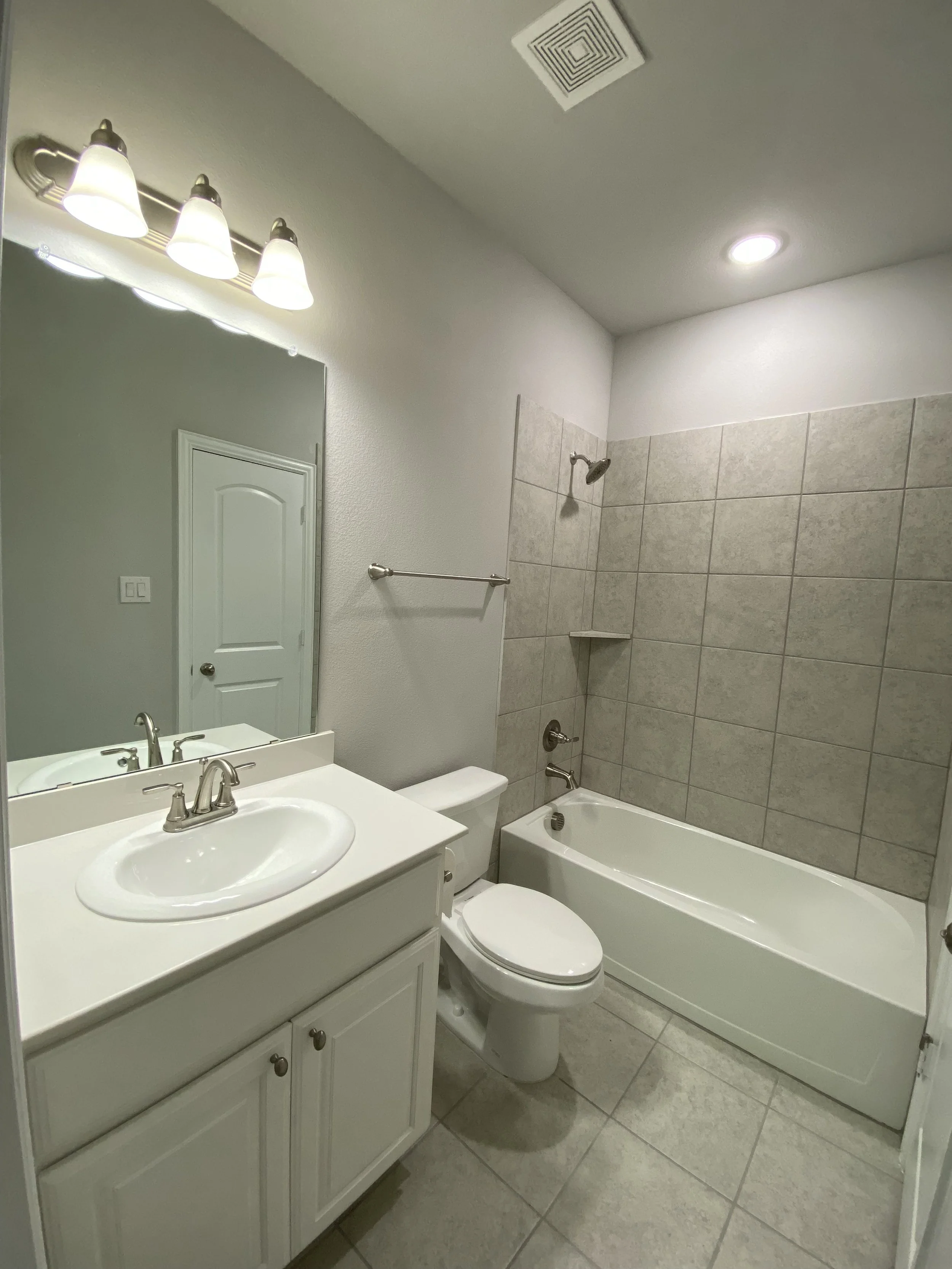 A small bathroom with a white vanity sink, a mirror, a toilet, a bathtub with beige tiles, a wall-mounted showerhead, a towel holder, a ceiling vent, and a ceiling light.