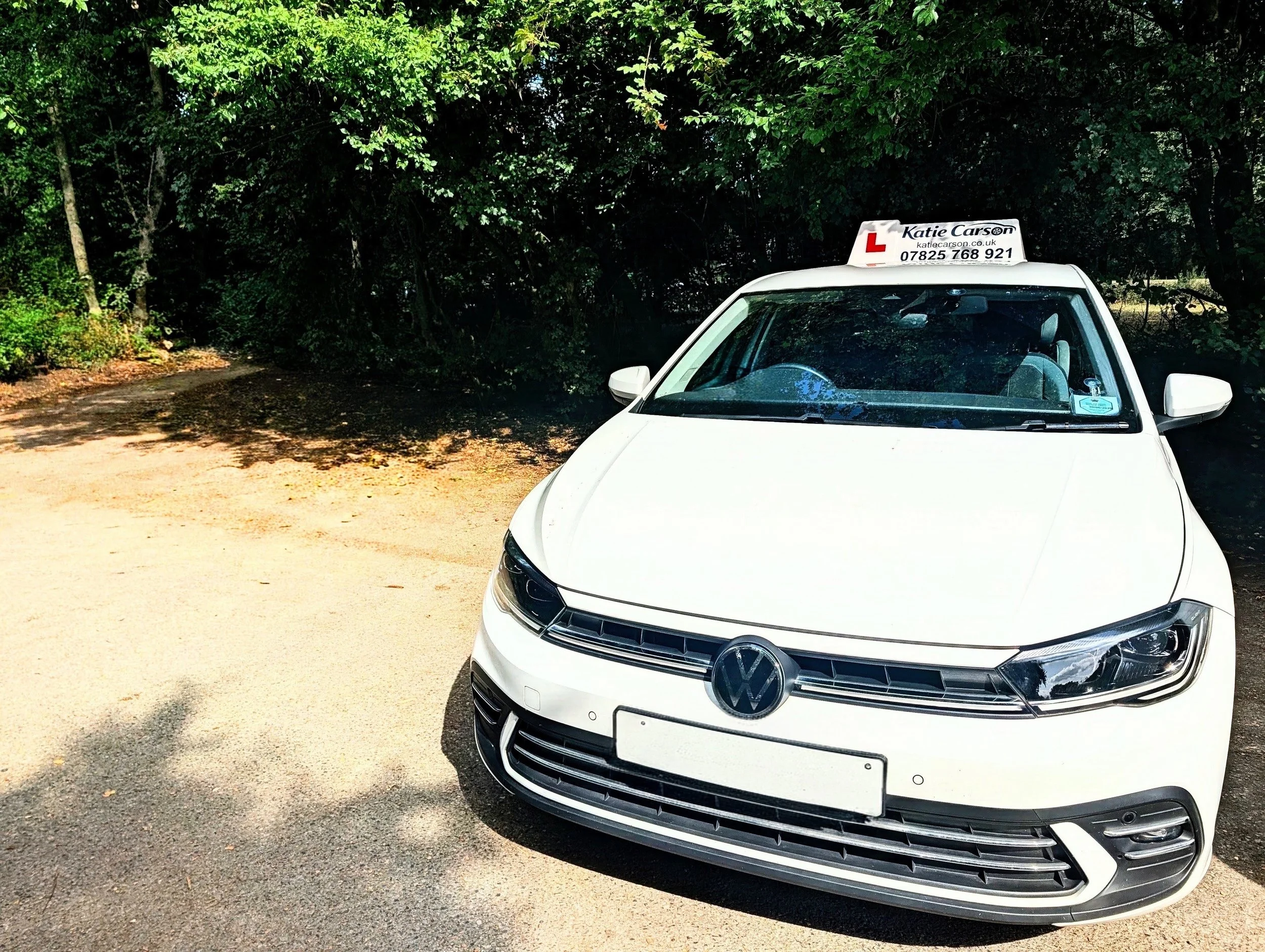 White Volkswagen car with driving instructor sign on top, parked on a gravel area near dense green trees.