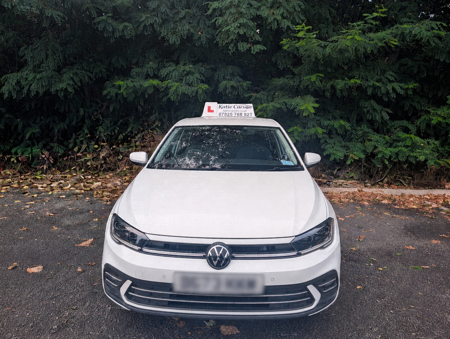 White Volkswagen car with driving instructor sign on top, parked on asphalt with fallen leaves, green trees and bushes in the background.