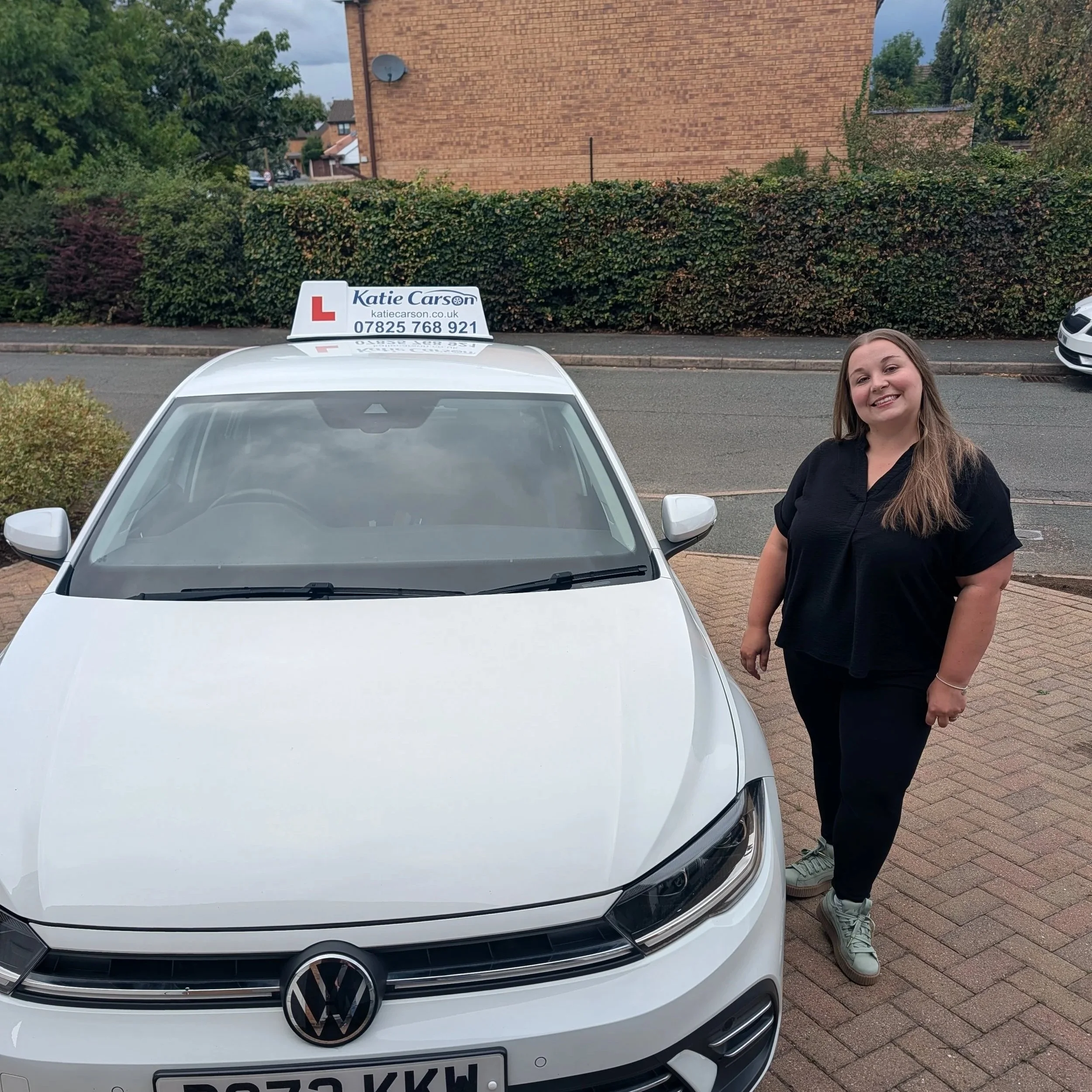 A white Volkswagen car parked on the street with a driver’s training sign on top that reads 'Katie Carson' with a large red 'L' and contact information, and a woman with long hair smiling standing beside it, in a suburban neighborhood with greenery, brick building, and cloudy sky in the background.