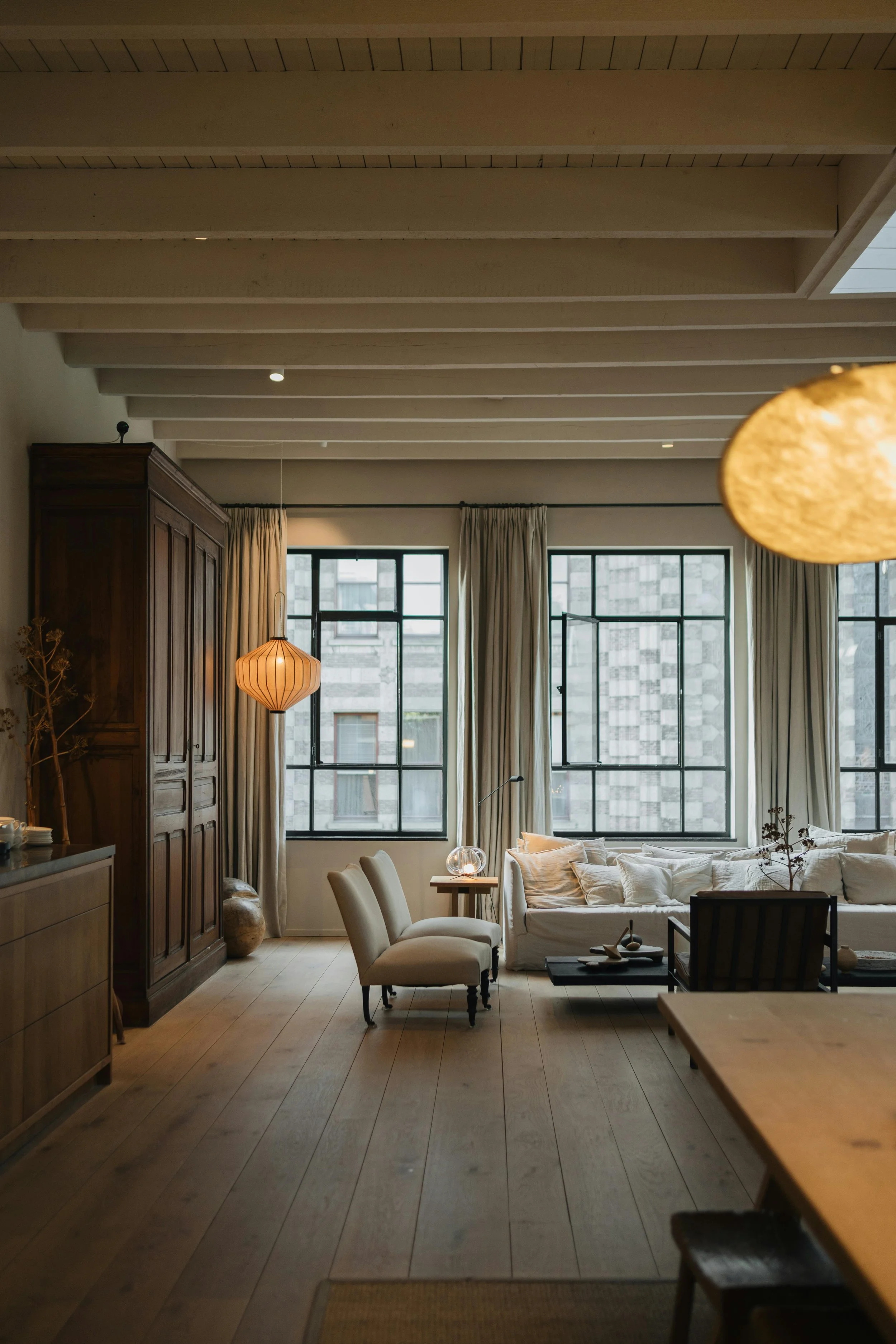 Living and dining space with exposed wood beam ceiling, dark wood built-ins, paper lantern pendant lights, and large windows with sheer curtains — Texas interior design project