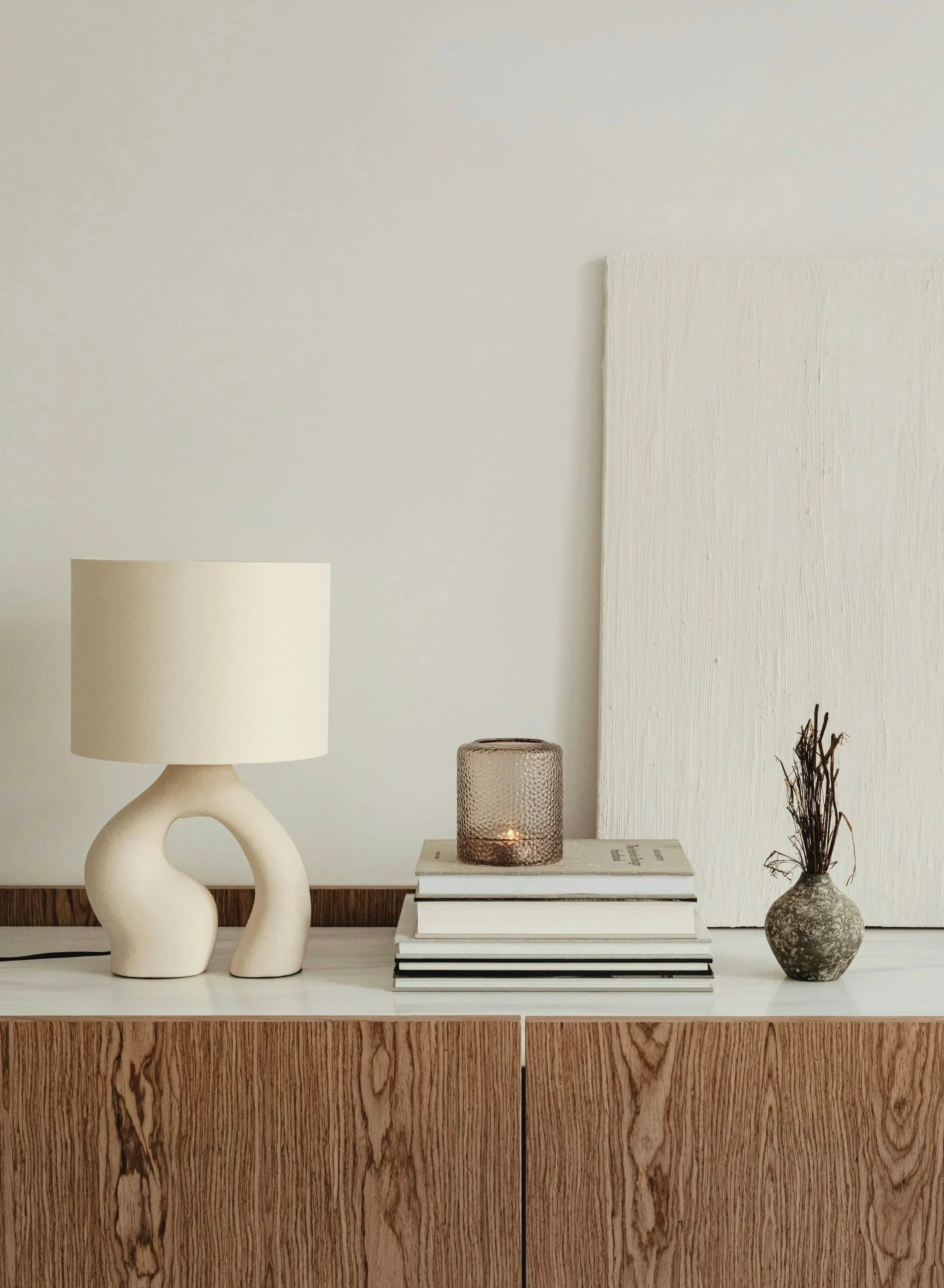 Wood credenza with a curved cream ceramic table lamp, stacked books, candle, and leaning framed art — interior styling detail