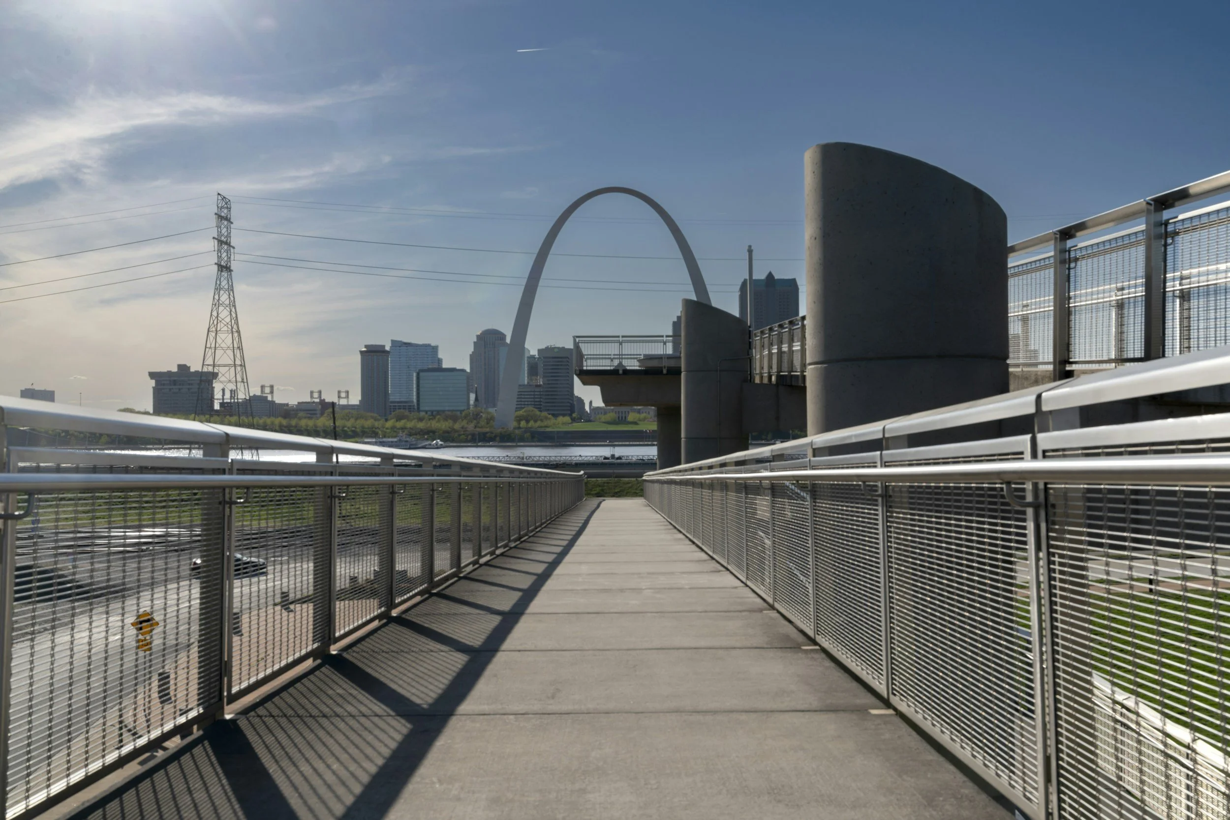 A pedestrian bridge in downtown St. Louis with the Gateway Arch in the background, high-rise buildings, a power line, and a bright blue sky. The bridge has metal railings and a wooden walkway, casting shadows.