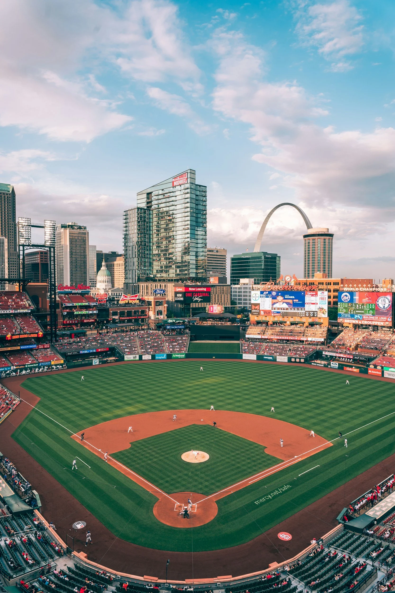 St. Louis Cardinals baseball field during game with players on the field and spectators in the stands, city skyline with Gateway Arch in the background.