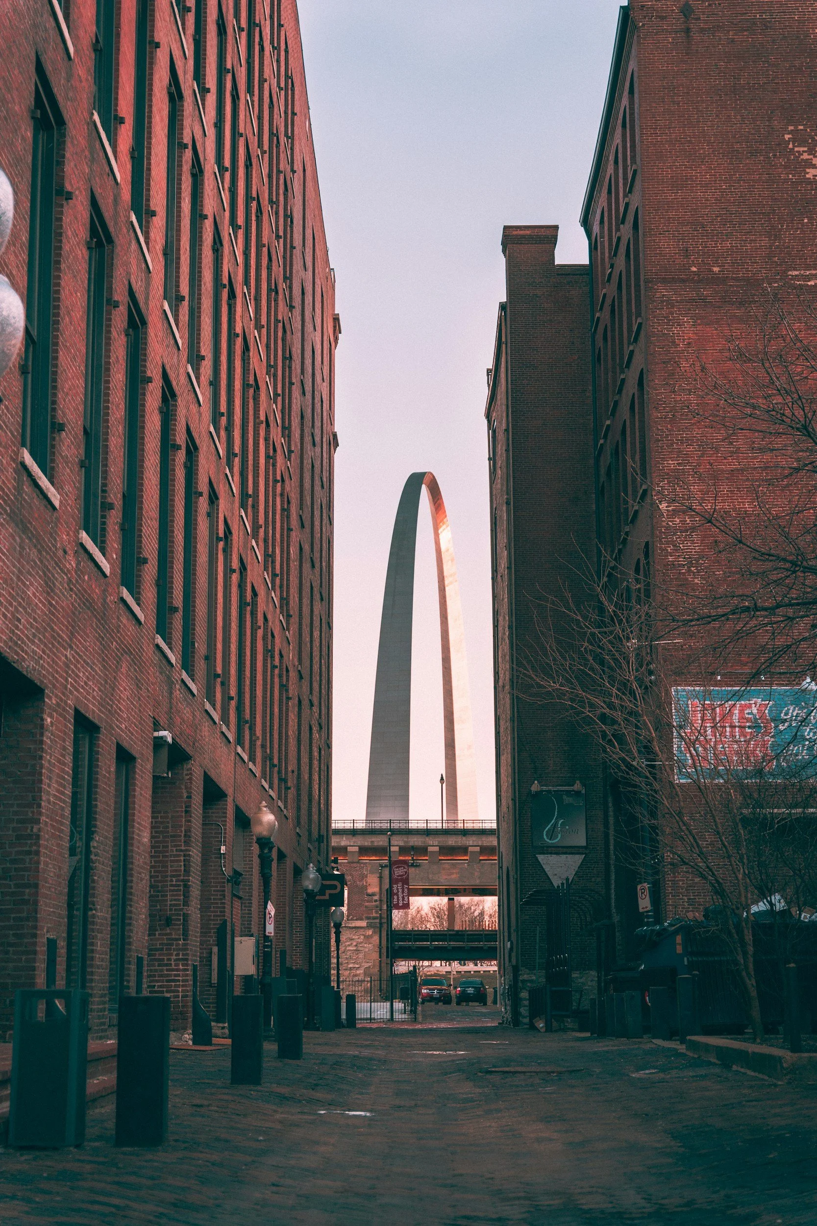 Looking down an urban alleyway flanked by brick buildings, with the Gateway Arch visible in the background.