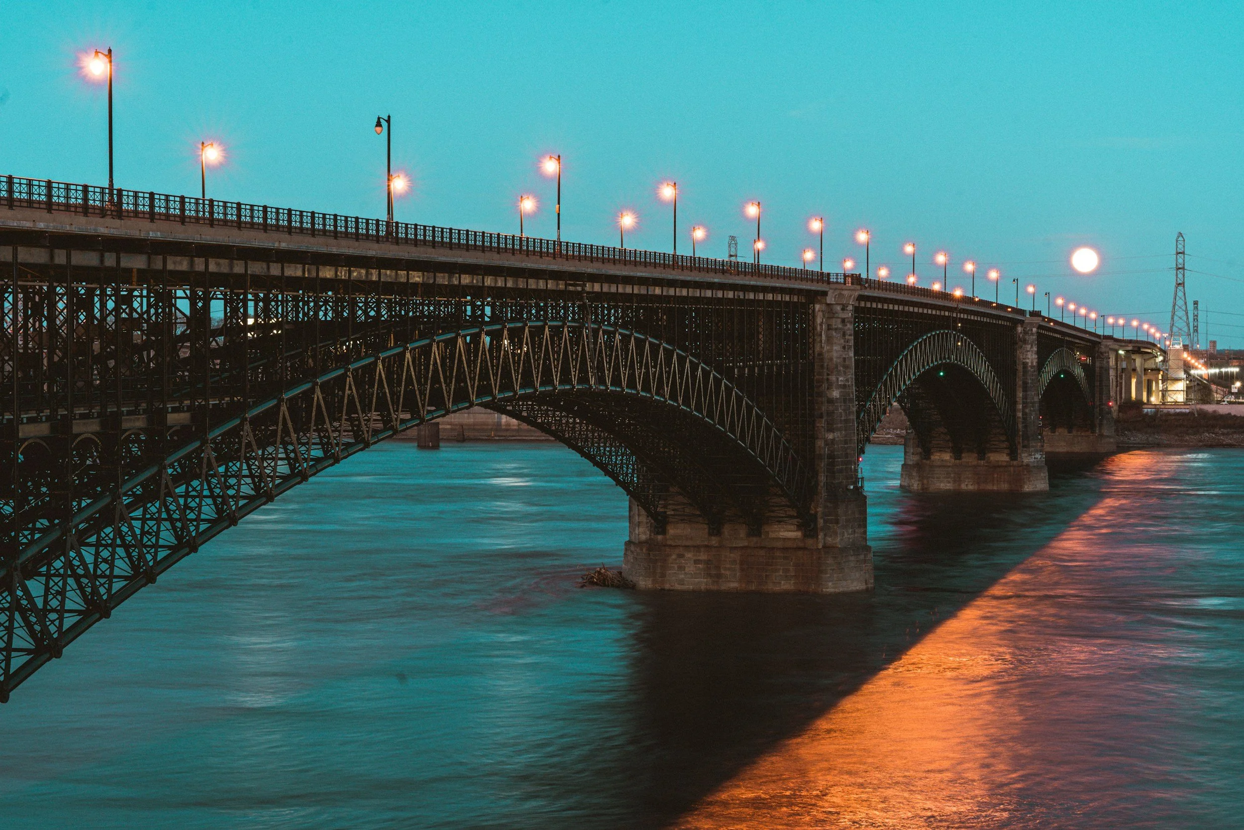 A steel arch bridge illuminated by streetlights over a body of water at dusk.