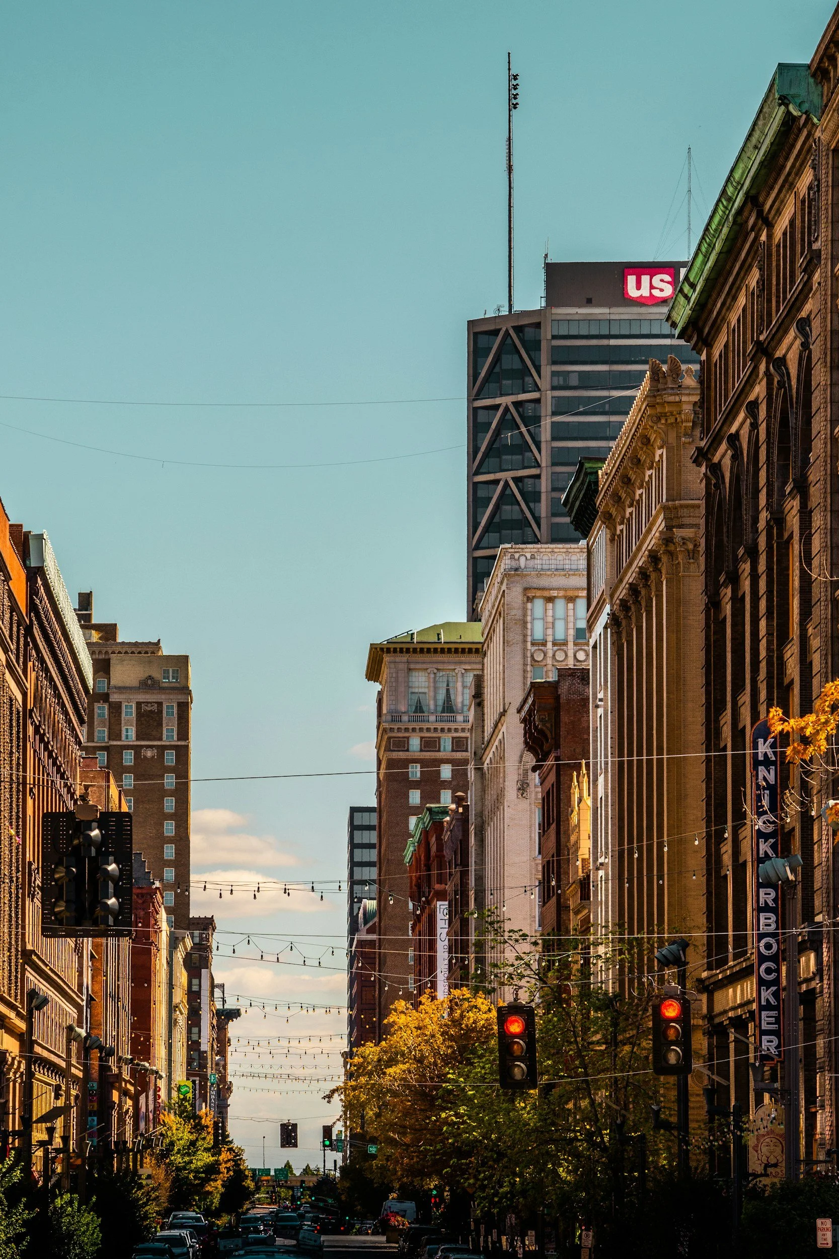 A city street with historic buildings on either side, trees, streetlights, and traffic signals, with a tall modern building with an 'US' logo in the background under a clear sky.